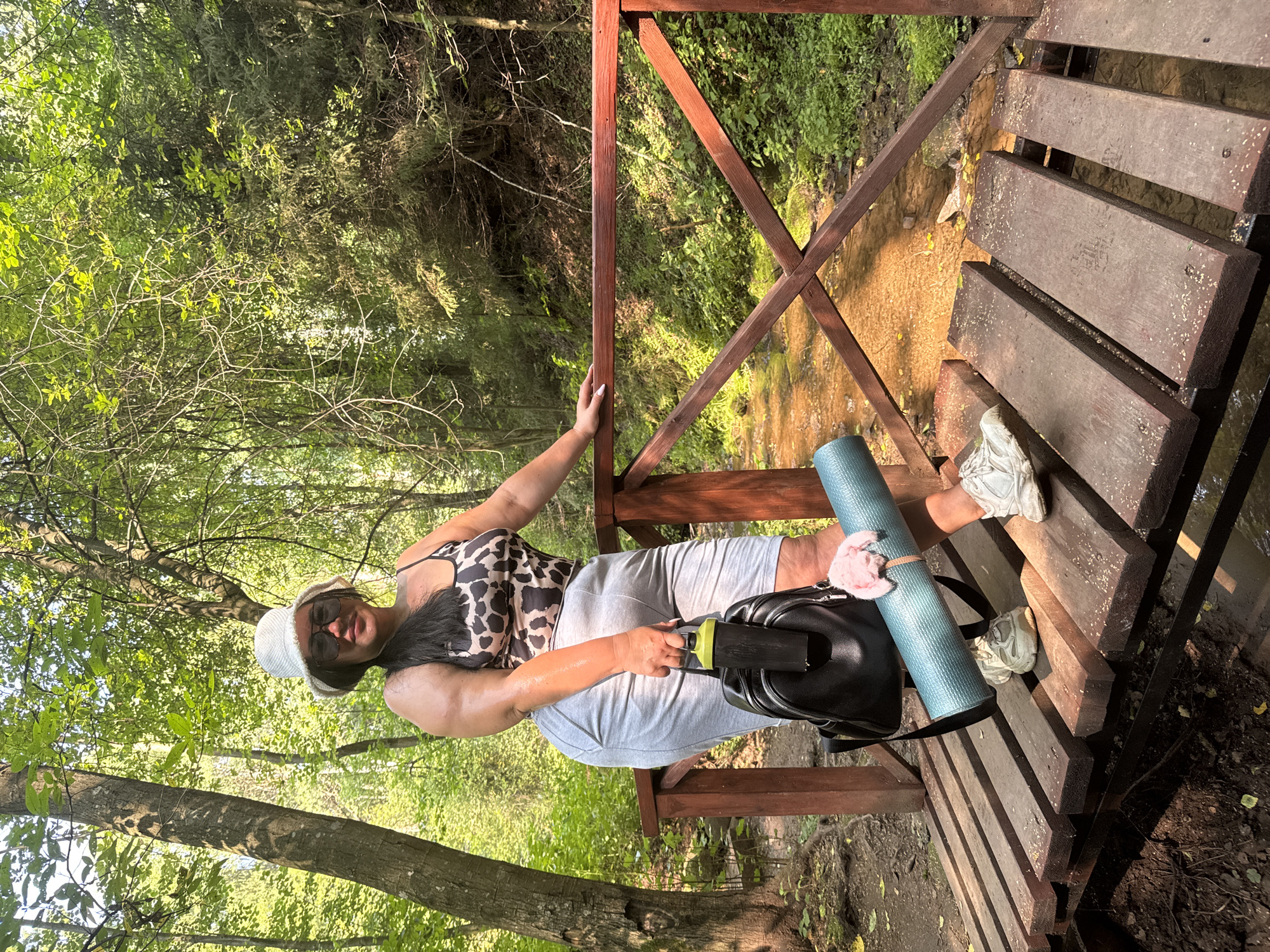 A full-body shot captures a person standing on a wooden bridge over a shallow stream in a forest. The individual is wearing a white bucket hat, dark sunglasses, a leopard-print tank top, and light gray shorts. They are holding a black backpack and a rolled-up teal yoga mat. The bridge has red wooden railings and planks. The surrounding environment is lush with green trees and foliage, with sunlight filtering through the canopy. The stream bed appears to be a mix of sand and pebbles. This scene is located in the park of the city of Izhevsk, Russia.