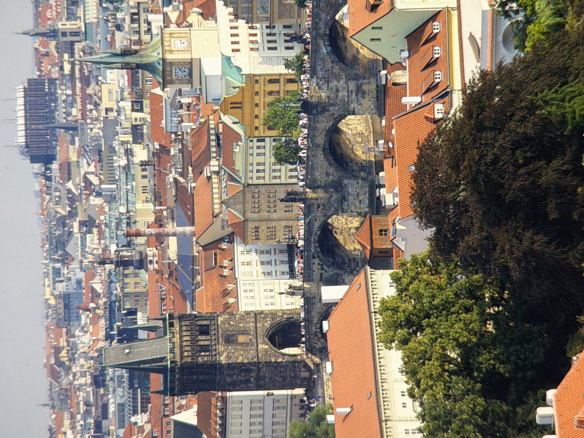 The image depicts a panoramic view of an old European city, characterized by its historic architecture, cobblestone bridge, and a skyline filled with numerous red-tiled roofs and spired buildings. In the foreground, a large, dark green tree with dense foliage partially obscures a cluster of buildings with terracotta roofs and dormer windows. Behind the tree, a stone bridge with multiple arches spans a body of water, with people visible walking across it. To the left of the bridge, a prominent, dark stone tower with a pointed roof rises above the surrounding structures. The background shows a vast expanse of buildings with varied architectural styles and colors, and a large, modern, striped building in the far distance. The sky is a clear, light gray. This scene is likely Prague, Czech Republic, specifically featuring the Charles Bridge and Old Town Bridge Tower, not Kyiv, Ukraine.