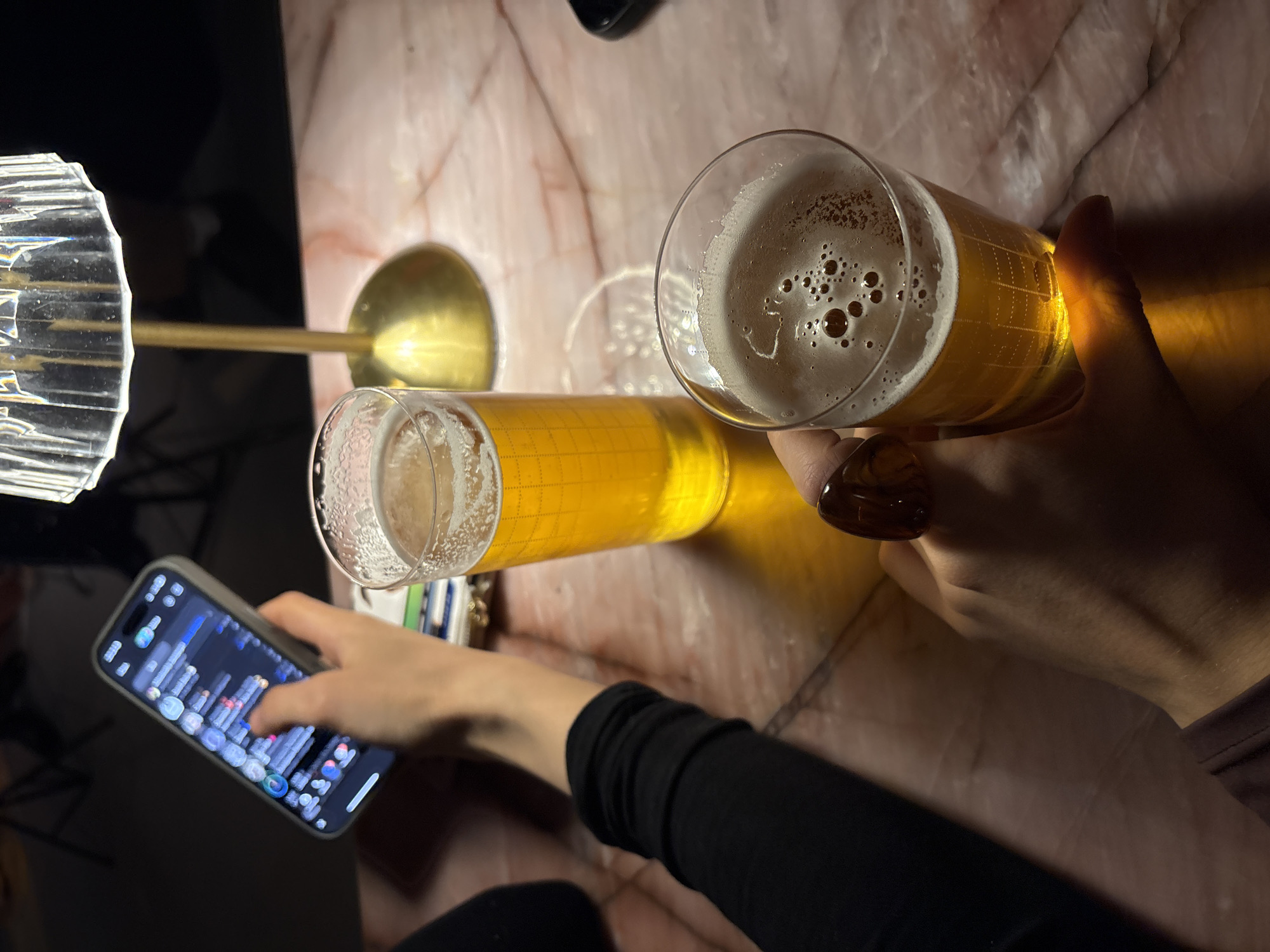 The image captures a moment at a table, possibly in a bar or restaurant. The table has a marble-like surface. Two glasses of beer are prominently featured; one is partially obscured by a hand adorned with a brown ring, and the other lies horizontally on the table. To the left, a hand is operating a smartphone displaying a financial graph. Adjacent to the horizontal beer glass are a pack of cigarettes and a lighter. The presence of a stylized lamp suggests the setting may be indoors. There is no evidence of the location being in Moscow, Russia.