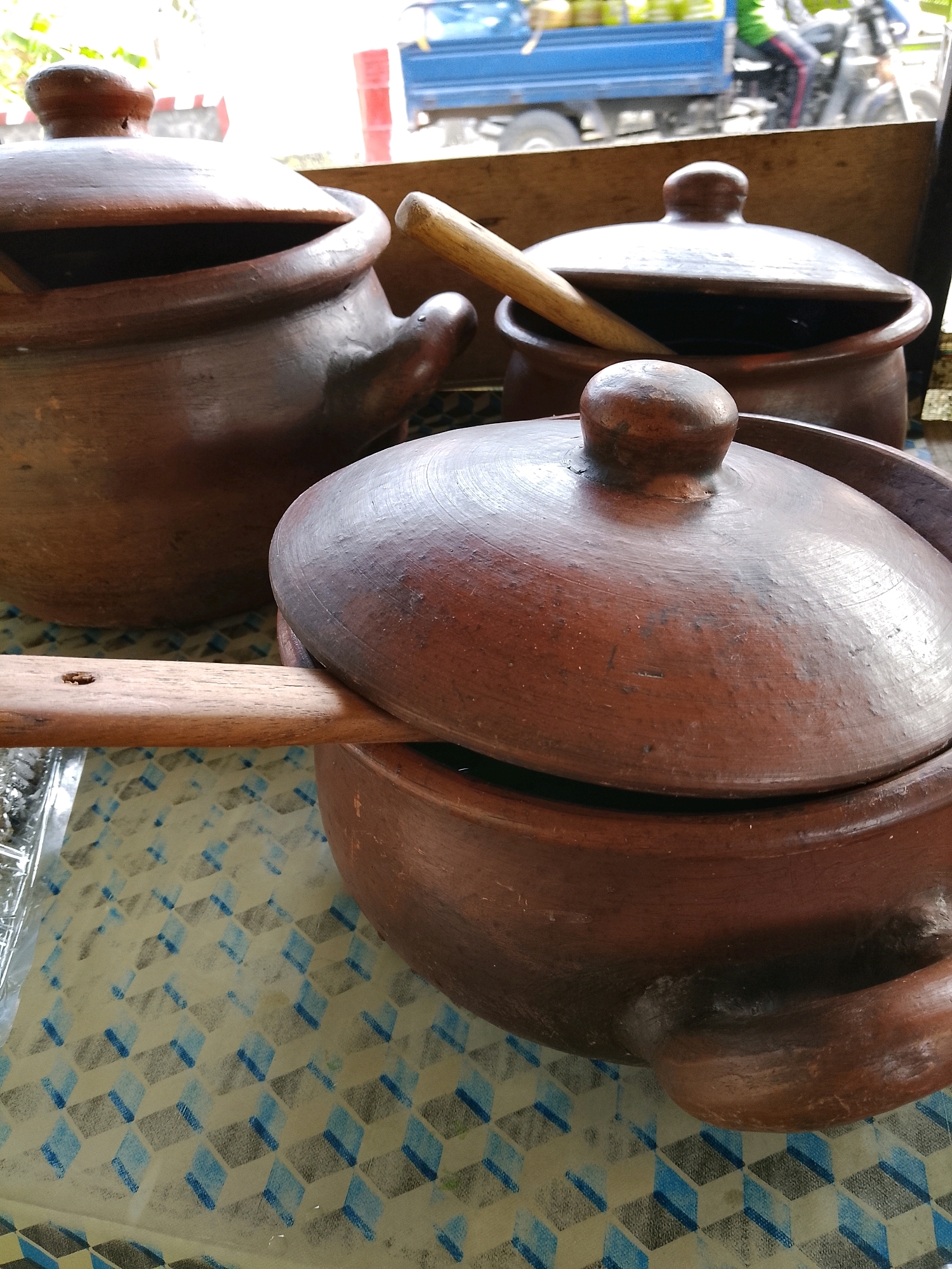 The image depicts a close-up, eye-level view of several terracotta clay pots with lids arranged on a table with a patterned tablecloth. The pots are of varying sizes, with two prominent ones in the foreground and a larger one partially visible in the background. A wooden ladle rests across the lid of the largest pot in the foreground, and another wooden utensil is positioned in one of the pots in the background. In the blurred background, a blue truck and a person on a motorcycle are visible, suggesting an outdoor setting, possibly a market or roadside stall in Banaran, Indonesia. The lighting suggests it is daytime, with natural light illuminating the scene. The tablecloth has a repeating geometric pattern of blue and gray squares.