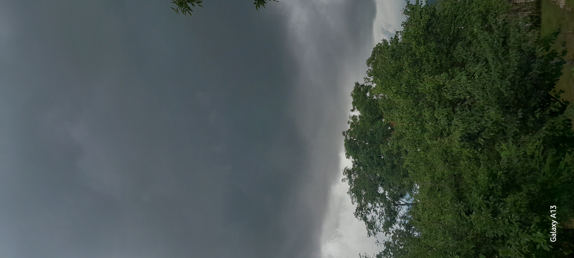 The image shows a view of dark, stormy clouds dominating the sky above a dense canopy of green trees. The foliage appears lush and full, indicating a temperate climate. In the lower left corner of the image, the text "Galaxy A13" is visible, suggesting this is a photograph taken with a Samsung Galaxy A13 phone. There are no people or animals present in the image, and no specific landmarks are identifiable. The environment appears to be natural, possibly a rural area or park.