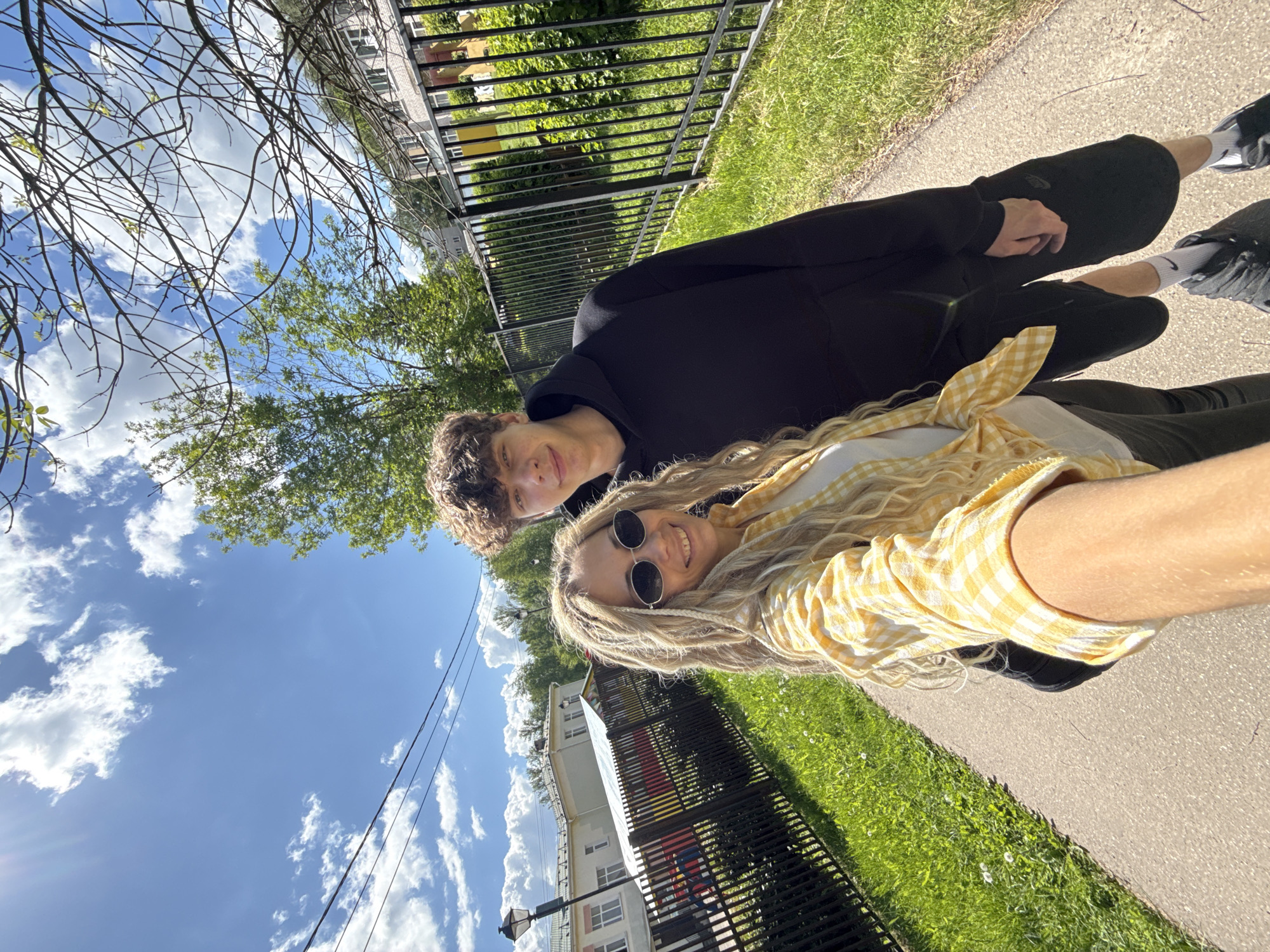 The image displays a cheerful outdoor selfie of two young individuals on a bright, sunny day in Rusanovo, Russia. The photo appears to be taken by the woman in the foreground, who has long, wavy blonde hair, round sunglasses, and is wearing a white top with a yellow and white checkered shirt tied around her waist. She is smiling broadly at the camera. Next to her, a man with curly dark hair and a black hooded sweatshirt also smiles.
They are positioned on what appears to be a paved path or sidewalk, with a strip of green grass and a black metal fence running parallel to them. Beyond the fence, a multi-story light-colored building with windows is visible, surrounded by lush green trees. The sky is a clear blue with scattered white, fluffy clouds, indicating pleasant weather. A street light pole is seen in the mid-ground, and thin power lines stretch across the upper part of the frame against the sky. The overall scene suggests a relaxed and happy moment enjoyed outdoors during daylight hours, likely in spring or summer. No visible text is present in the image.