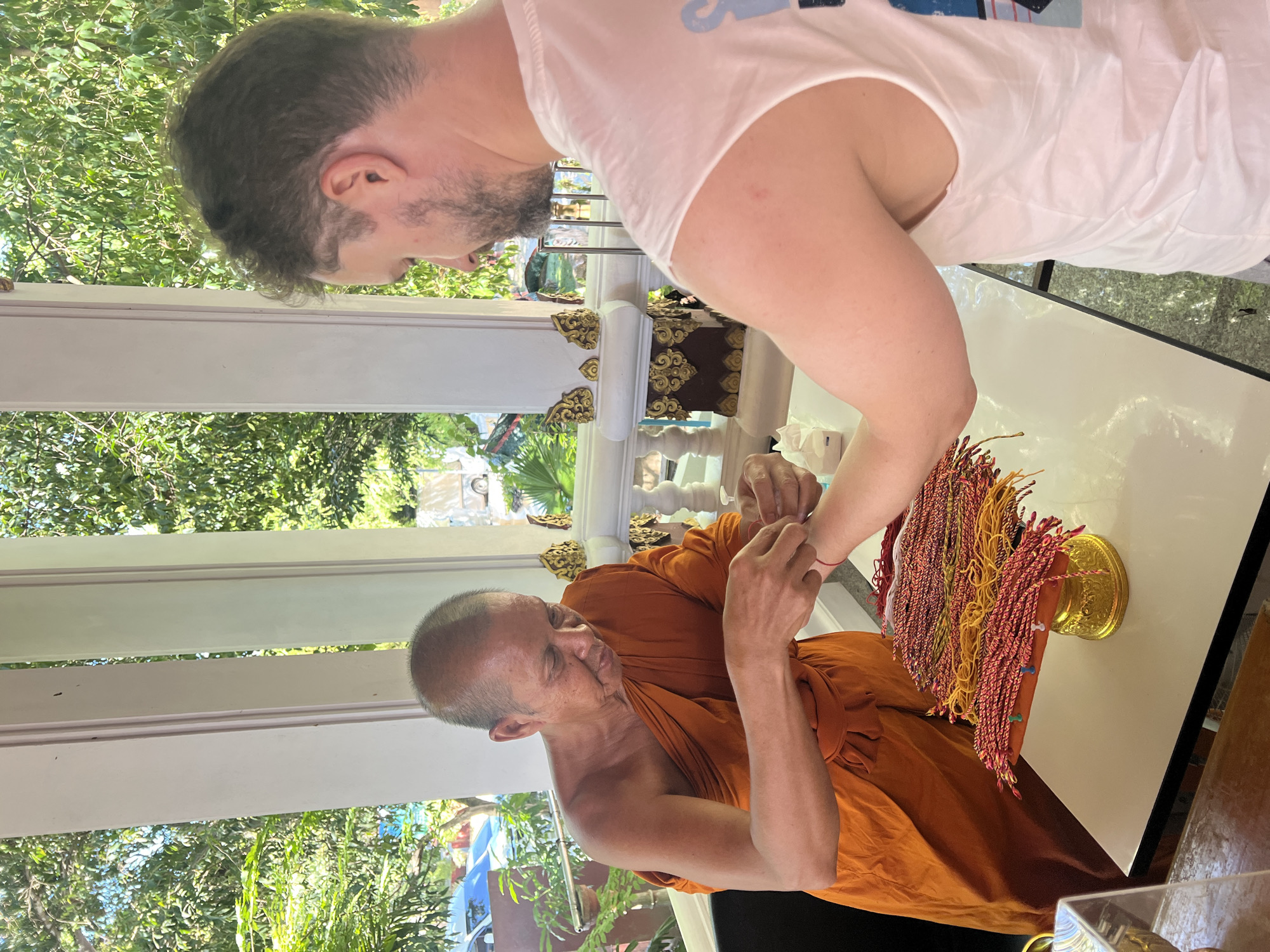 In Baan Hua Thanon, Thailand, a Buddhist monk dressed in an orange robe is seen tying a red string bracelet onto the wrist of a man with short brown hair and a beard. The man is wearing a white tank top. They are standing at a table, where a golden tray with numerous colorful braided string bracelets, primarily red and yellow, is placed. In the background, white pillars with ornate golden decorations are visible, along with lush green foliage and trees, suggesting an outdoor or semi-outdoor setting. The lighting indicates it is daytime, likely under sunlight.
