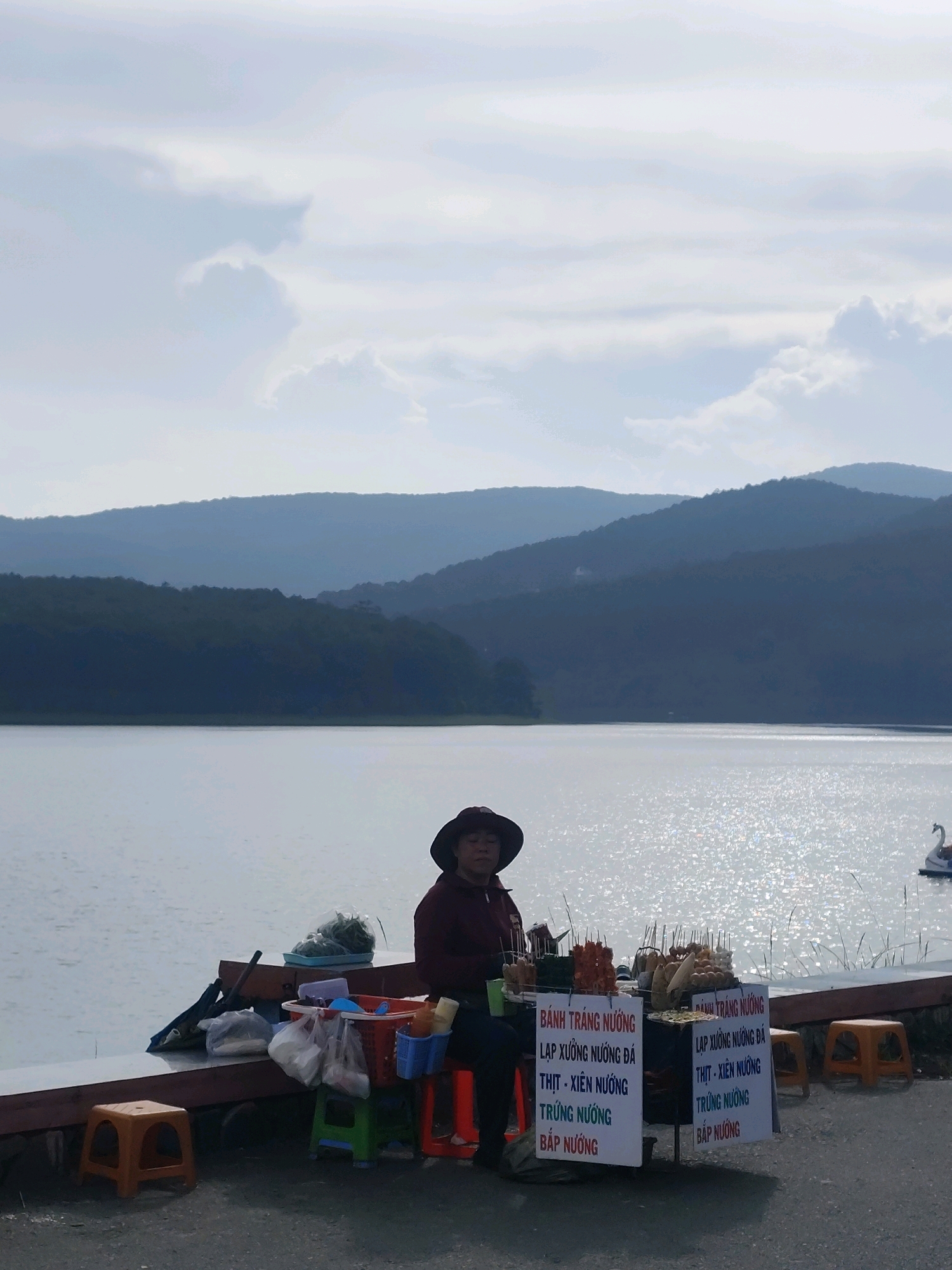 A woman wearing a wide-brimmed hat and a maroon top sits beside a food cart at the edge of a lake, likely Xuan Huong Lake in Da Lat, Vietnam. The food cart displays skewers of grilled meat and corn, with signs written in Vietnamese advertising "Banh Trang Nuong" (grilled rice paper) and various grilled items like "Lap Xuong Nuong Da" (grilled sausages), "Thit - Xien Nuong" (meat skewers), "Trung Nuong" (grilled eggs), and "Bap Nuong" (grilled corn). Bags of plastic wrap, a basket of vegetables, and a few plastic stools are also visible around the cart and on the adjacent walkway. The lake is calm and reflects the bright sunlight, with distant, hazy blue mountains forming the backdrop under a cloudy sky. A white swan-shaped paddle boat is visible in the water to the right.