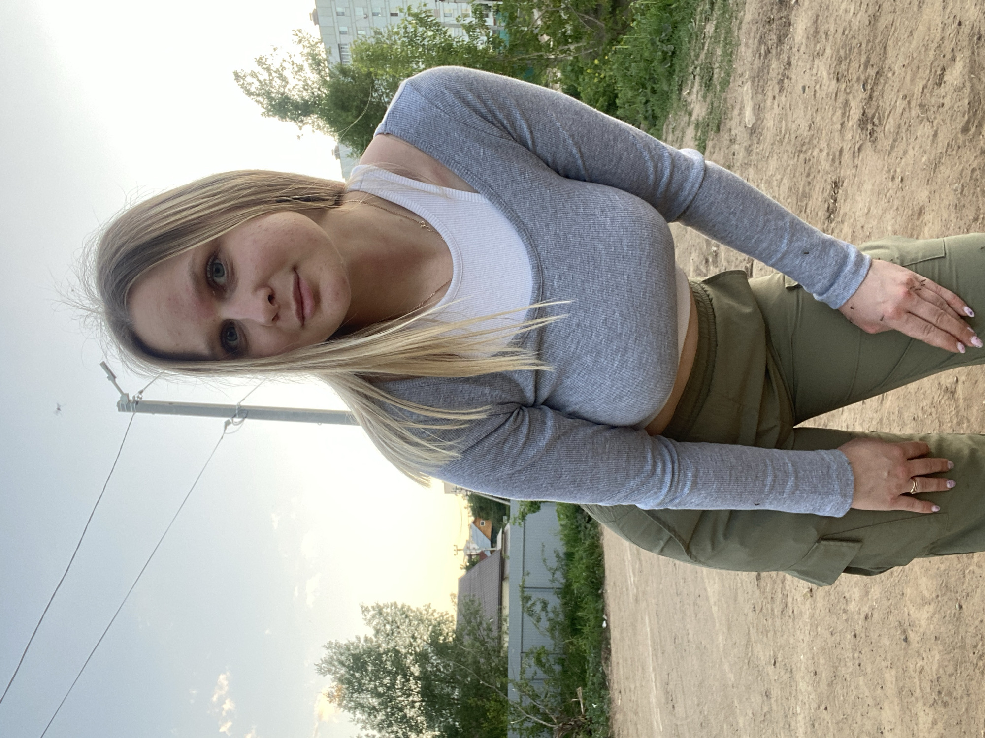 A young woman with blond hair, wearing a grey long-sleeved shirt over a white top and green cargo pants, is positioned outdoors. The location is inferred to be Chaikovsky, Russia, based on the request context. The background shows a mix of urban and natural elements, including buildings, trees, and a dirt or gravel path. Overhead power lines are visible. The lighting suggests it is daytime.