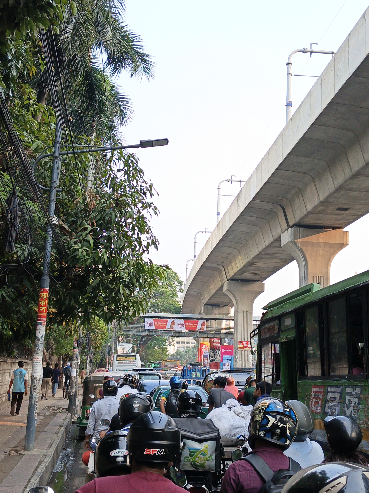 The image shows a street scene in Dhaka, Bangladesh, characterized by heavy traffic congestion and urban infrastructure. The foreground is dominated by a dense cluster of motorcycles and auto-rickshaws, mostly occupied by helmeted riders and passengers. On the left, pedestrians walk along a narrow sidewalk adjacent to the traffic. A concrete elevated railway structure looms overhead, with vertical support pillars visible amidst the vehicles. Street signs, banners, and advertisements are affixed to the overpass structure. Lush, green vegetation, including a tall palm tree and other trees, partially obscures buildings and infrastructure on the left side of the frame. Utility poles laden with tangled wires are also present. In the middle distance, more vehicles are visible, indicating a traffic jam. The image captures the frenetic urban atmosphere typical of Dhaka.