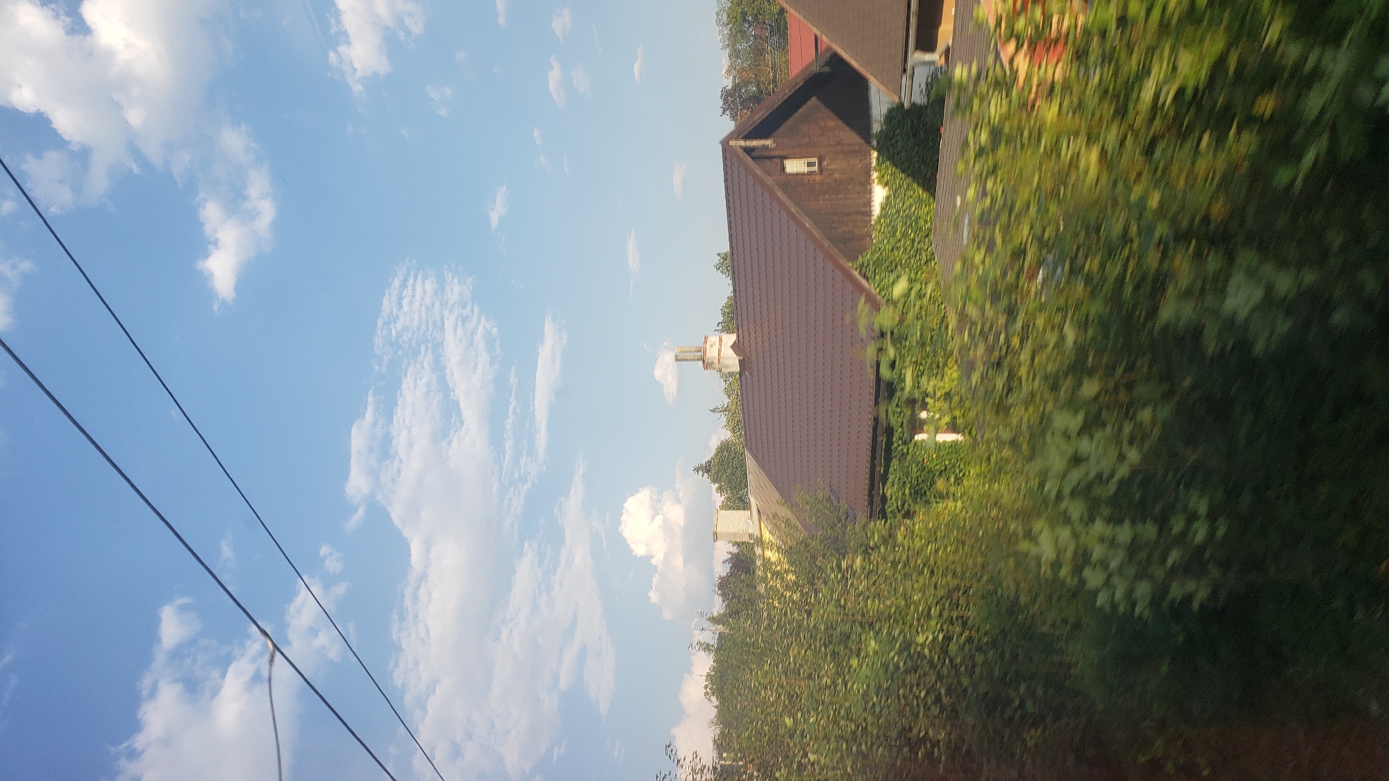 The image depicts a view of rooftops and vegetation, likely taken from a moving vehicle, suggesting a roadside or train view. In the foreground, lush green bushes and trees are blurred due to motion, partially obscuring the view of buildings behind them. Several houses with brown, tiled roofs are visible. One prominent house has a dark wood exterior and a large, steep brown roof with a small, square window. Another house to its left has a lighter colored exterior and a similarly tiled roof. Two chimneys are visible; one is a tall, cylindrical structure with visible brickwork, and another is shorter and more integrated with a roofline. In the background, more trees are visible, and the sky is a clear blue with scattered white clouds. The presence of power lines in the upper part of the image further suggests an urban or suburban setting. While the image provides a general view of houses and greenery, there are no visible people or specific landmarks that would allow for precise identification of the location within Ružomberok, Slovakia, without additional context or metadata.