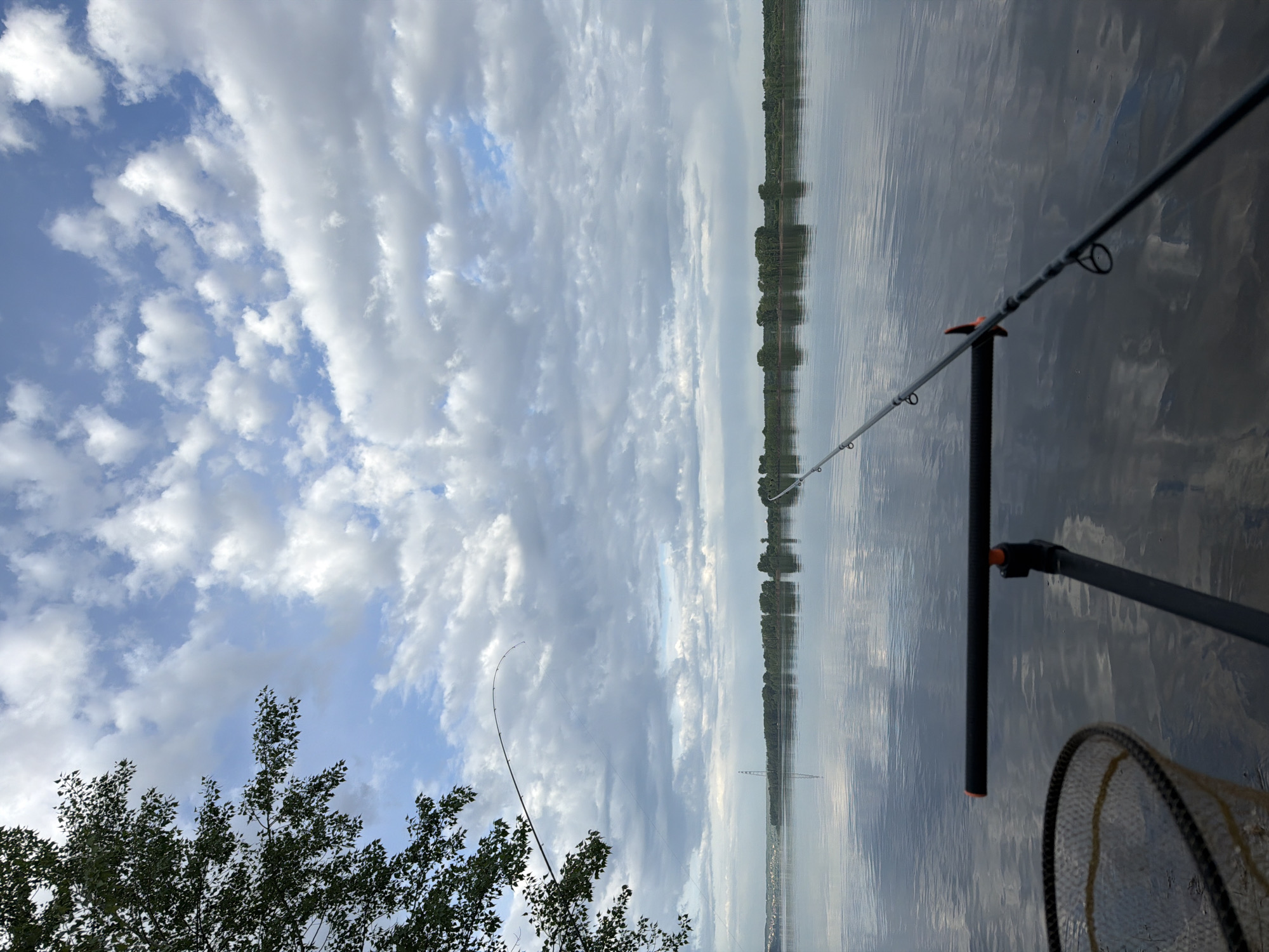 The image depicts a fishing scene, likely on or near the Kama River, in Chaikovsky, Russia. A fishing rod is visible in the foreground, with its line extending toward the water. A fishing net is also present, suggesting the activity of catching fish. The background shows a body of water reflecting the sky, which is filled with clouds. The shoreline is visible in the distance, lined with trees and foliage. Some trees can be seen in the foreground on the left-hand side of the image. The scene is captured during daylight, with natural lighting and an outdoor environment.