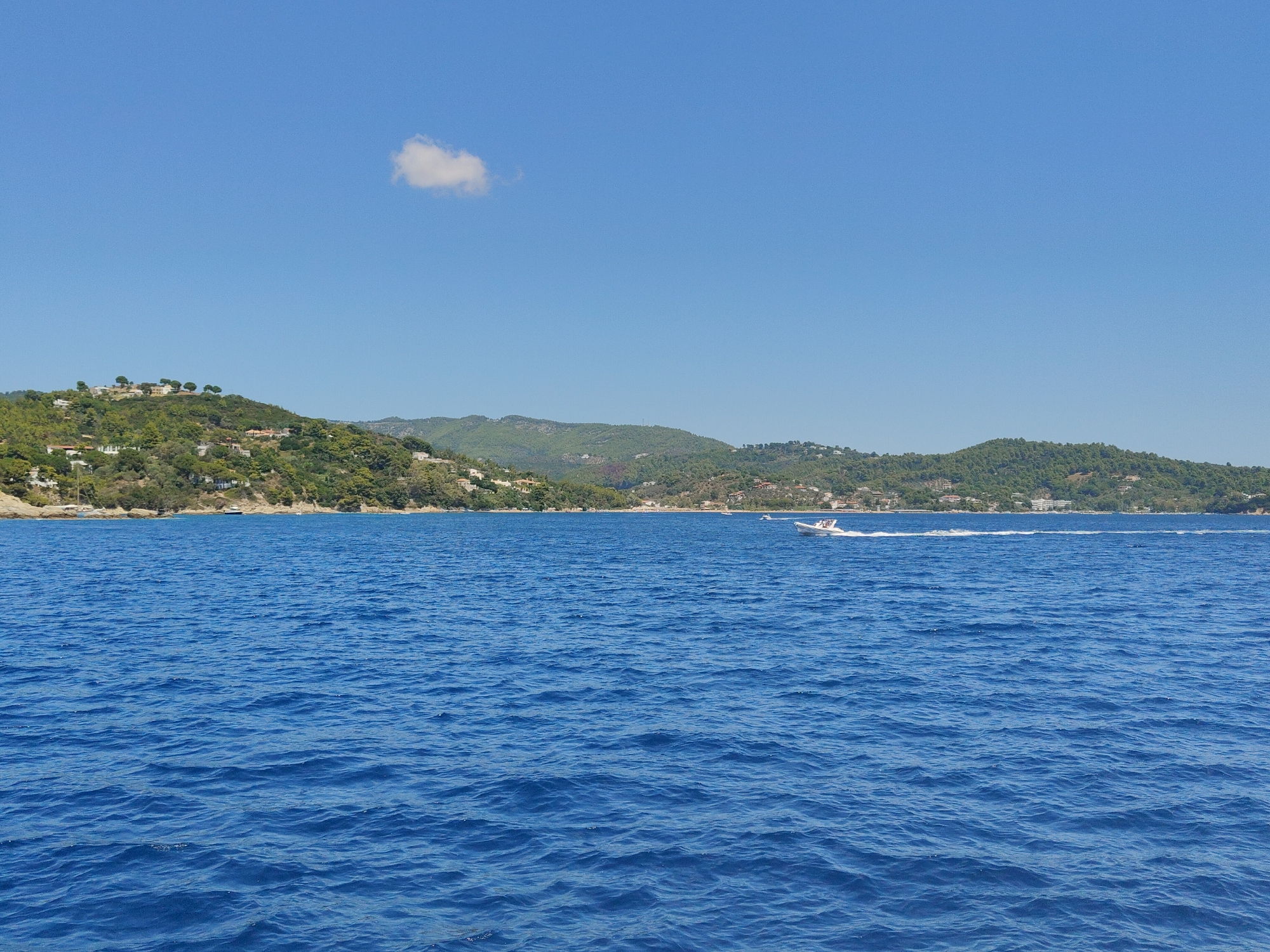 The image depicts a vibrant coastal scene under a clear blue sky with a single, fluffy white cloud. The foreground is dominated by the deep blue, rippling waters of the sea. In the mid-ground, a coastline rises, densely covered in lush green trees and dotted with white buildings, suggesting a populated area. A small, white motorboat with people aboard is speeding across the water, leaving a white wake behind it. The background features rolling, verdant hills stretching towards the horizon. While the specific location of Platanias, Greece, is not explicitly visible, the scenery is consistent with a Mediterranean coastal town.