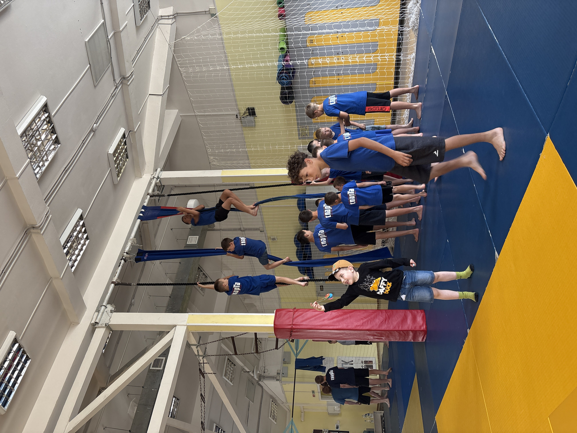 The image depicts an indoor gymnastics facility, likely a children's sports club, located in Chaikovsky, Russia. Several young boys, all wearing blue t-shirts, are engaged in various activities. Some are actively climbing ropes suspended from the ceiling, while others are observing. One boy in the foreground, wearing a black long-sleeved shirt and denim shorts, is holding a yo-yo and appears to be showing it off to the other children. The environment includes gymnasium mats on the floor, safety nets along the wall, and fluorescent lighting fixtures on the ceiling. The overall atmosphere suggests a training session or a recreational activity for young athletes.