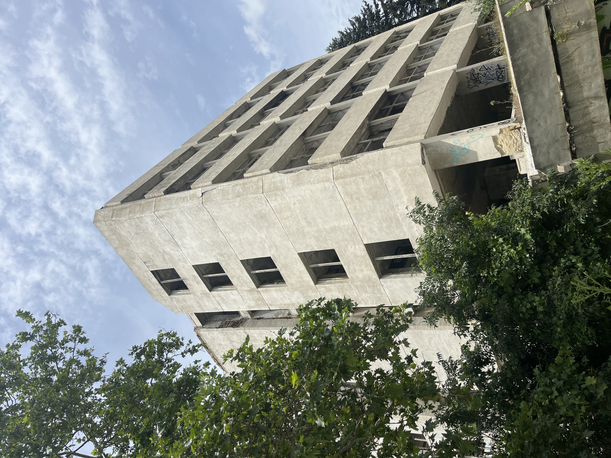 The photograph depicts a large, abandoned, multi-story concrete building, viewed from a low angle against a partly cloudy sky. The building is in a state of severe disrepair, with many windows missing and visible structural damage, including cracks and exposed rebar on the exterior. Graffiti is present on some of the lower concrete surfaces. Lush green trees and foliage surround the lower sections of the building, partially obscuring it. A dark evergreen tree is visible to the right of the concrete structure. The scene suggests an urban environment that has experienced decay or neglect. This building is the former "Communist Party House" in Varna, Bulgaria, a structure with a controversial history and a prominent feature of the city's skyline, often referred to as the "Palace of the Republic" or "Dom na Partiyata".
