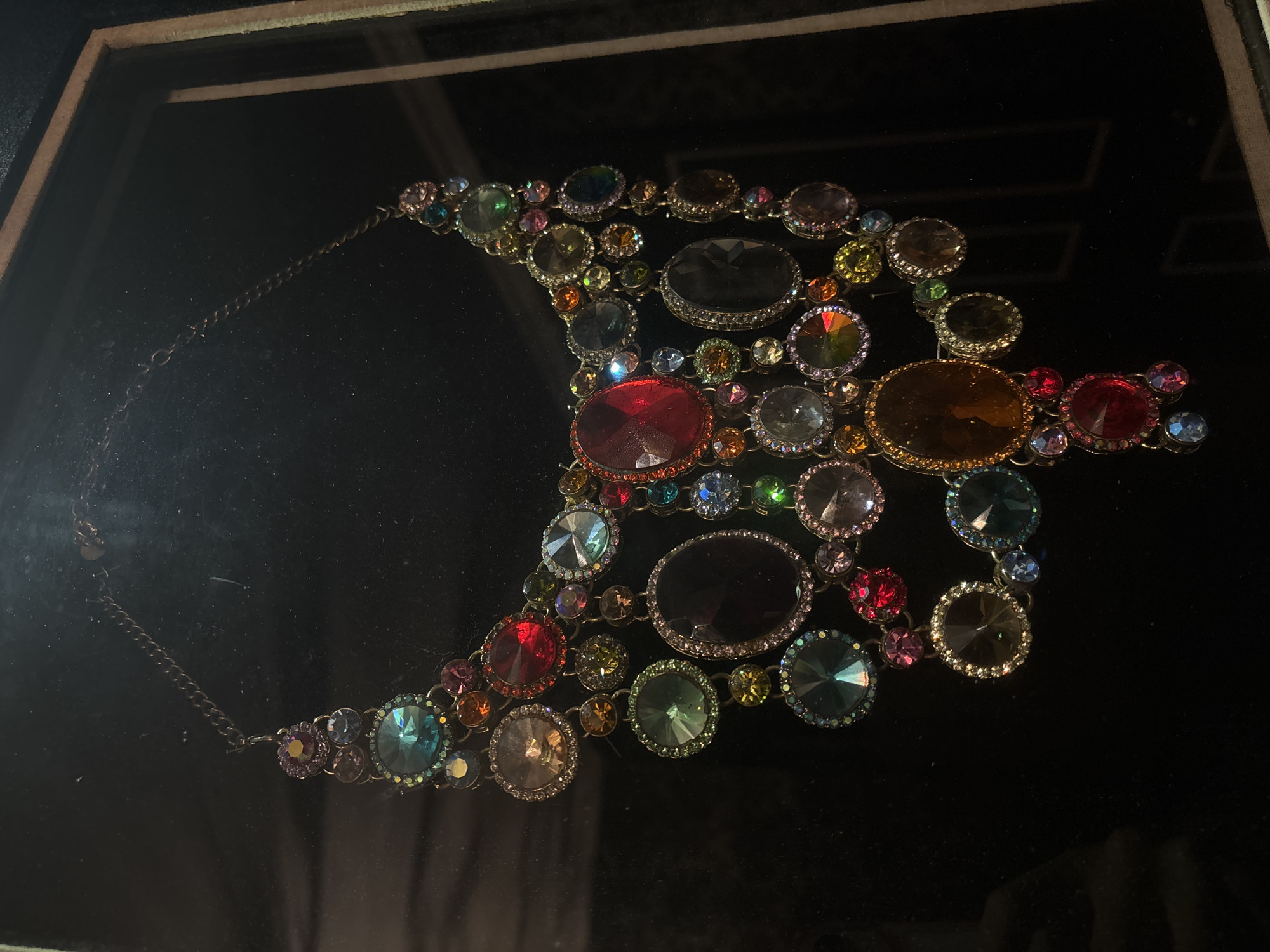 The image shows a necklace with multiple large, colorful gemstones set in metal, displayed against a dark, velvet-like background within a glass-covered frame. The necklace is a crescent shape, with a chain extending from one end. The gemstones are various sizes and colors, including red, green, blue, amber, and clear, each encircled by smaller, clear stones. There is a dark, oval-shaped gem in the necklace. The setting seems to be a formal display case. There is no indication of this image being from North Kuta, Indonesia.