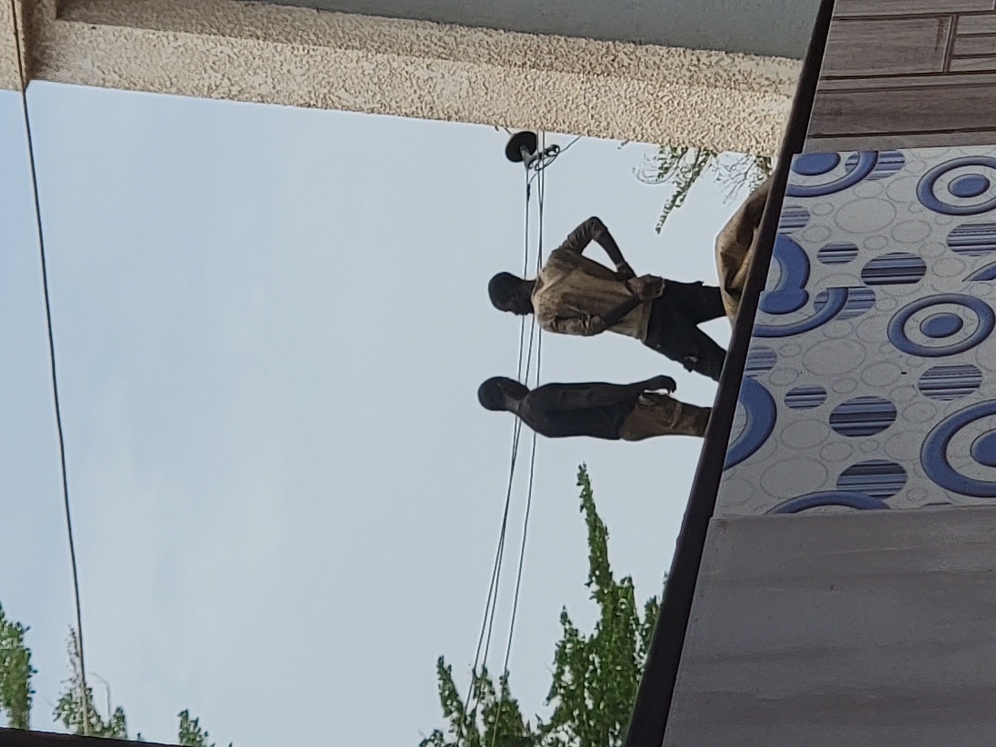 Two men are observed working on overhead power lines in Maiburiji, Nigeria. One man is wearing a light-colored shirt and dark pants, while the other is wearing a dark shirt and light-colored pants. They are positioned between a building with a textured exterior and another with patterned tiles. Green foliage is visible in the lower portion of the image.
