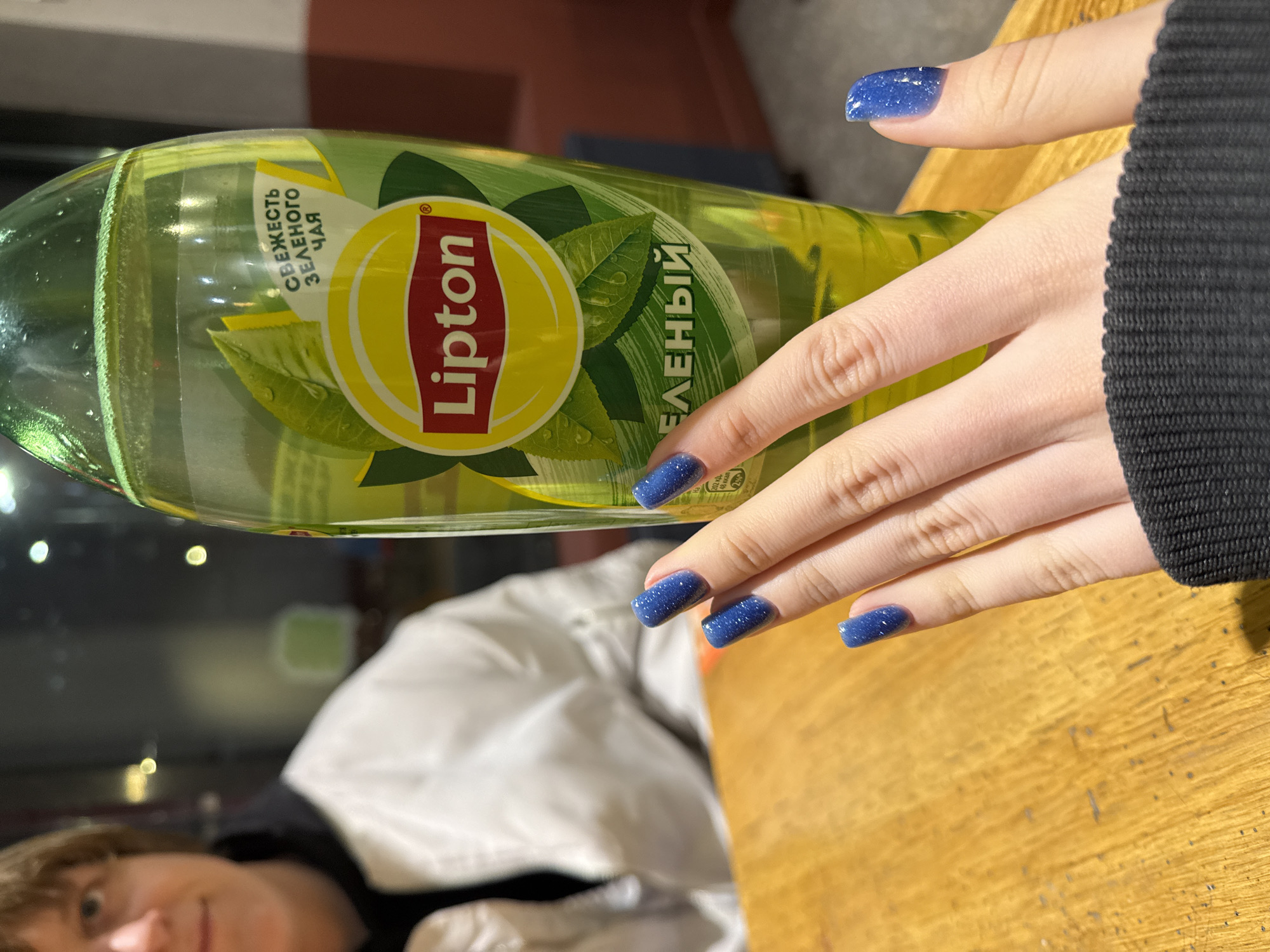 A close-up shot presents a bottle of Lipton green tea against a blurred background. The bottle's label is visible, featuring the Lipton logo and text in Russian. A person's hand is positioned near the bottle, showcasing nails painted in a dark blue color. The background includes a person in a white jacket and an indistinct cityscape. Given the Russian text on the bottle and general appearance, the location is likely Saint Petersburg, Russia. The scene captures a moment of consumption or display.
