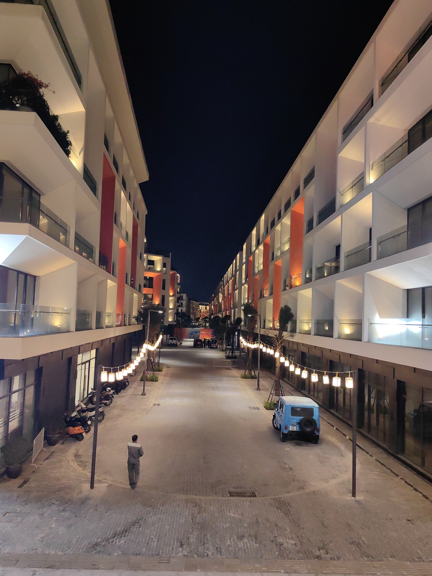 The image shows a street scene at night in Bai Chay Ward, Vietnam. Two modern, multi-story buildings with white facades and orange accent columns flank a brick-paved road. String lights hang between the buildings along the road's edge. A man in work clothes walks away from the viewer, toward the center of the street. A light blue, boxy vehicle is parked on the right side of the street. Motorcycles are parked along the left side of the street. The sky is dark.