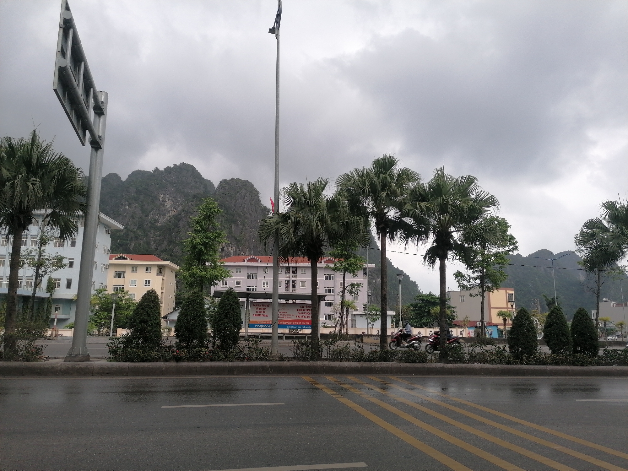 This image presents an overcast street scene in Ha Tu Ward, Vietnam, characterized by a wide, wet asphalt road in the foreground, suggesting recent rain. Yellow diagonal markings are visible on the road surface. A central median strip features neatly trimmed bushes, small evergreen trees, and several tall palm trees.
Beyond the median, a row of multi-story buildings lines the street, including a light blue structure on the left, a light yellow building further back, and a prominent greyish-white building with a red tiled roof. A large, metallic street sign or billboard frame is visible on the left side, and a very tall, slender pole extends into the cloudy sky from the center.
In the middle ground, two individuals on motorcycles are seen riding from left to right on the far side of the road, indicating everyday urban activity. Majestic karstic mountains, partially covered in greenery, dominate the background under a heavy, grey, cloudy sky.
A large billboard in front of the grey building displays Vietnamese text. Visible phrases include "XĂM HẠI PHỤ NỮ VÀ TRẺ EM LÀ TỘI ÁC NGHIÊM TRỌNG" (Sexual abuse of women and children is a serious crime) and "HÃY TỐ CÁO HÀNH VI XÂM HẠI BẠO LỰC ĐỐI VỚI PHỤ NỮ VÀ TRẺ EM" (Report acts of abuse, violence against women and children). The billboard also mentions "vinaphone" with a slogan that appears to be "SIÊU TỐC ĐỘ SIÊU TRẢI NGHIỆM" (Super speed, super experience). The daytime scene has a muted quality due to the heavy cloud cover.