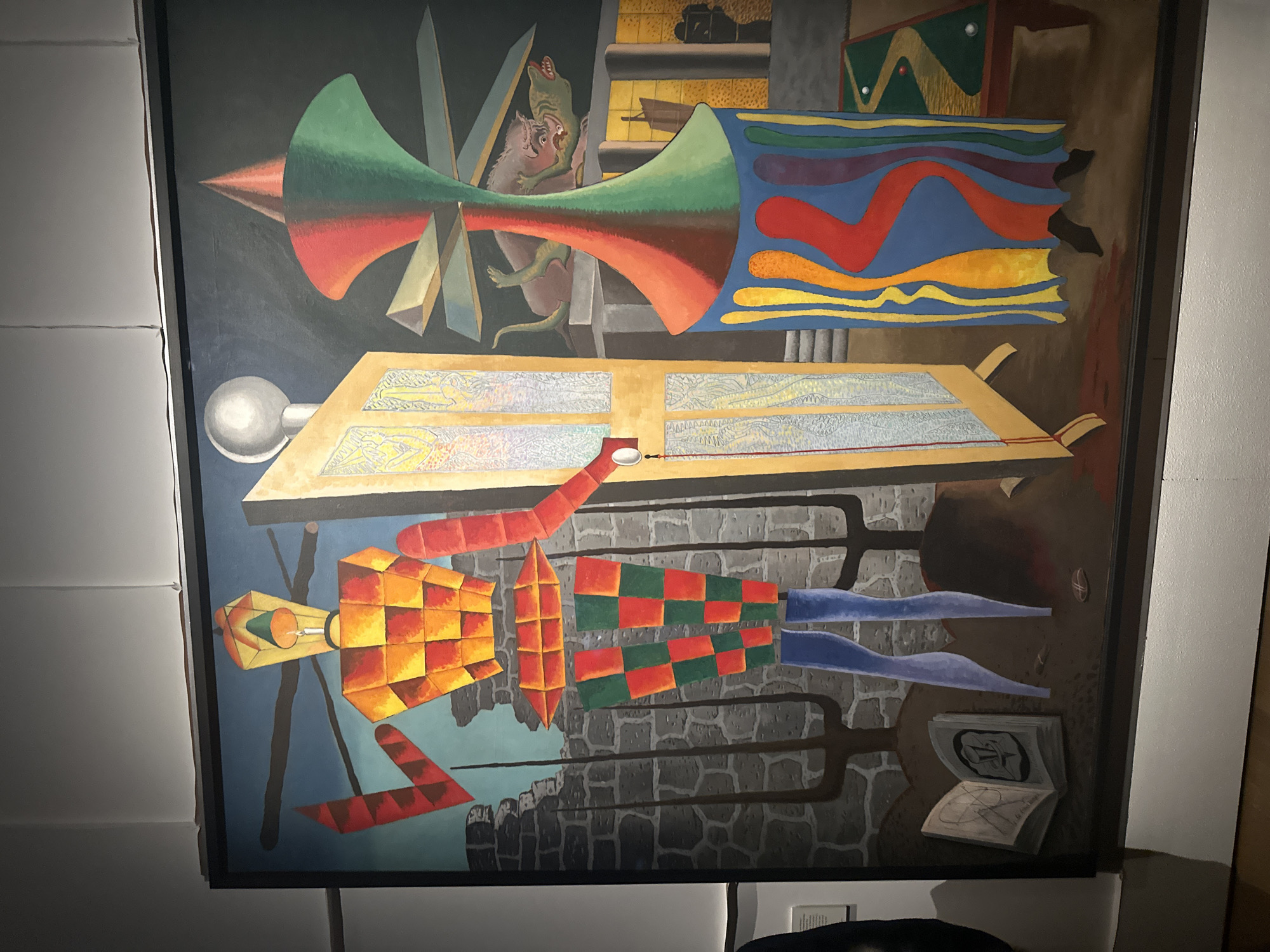 The image displays a large, framed oil painting titled "The Bride" by Max Ernst, exhibited at the Tate Modern in London, United Kingdom. The surrealist artwork features a stylized, geometrically segmented humanoid figure on the left, primarily constructed from orange and yellow checkered blocks for the torso and head, red angular arms, and red and green checkered blocks for the upper legs, with solid blue lower legs. Its head resembles a yellow lantern with an internal light source. The figure's right arm extends towards a tall, narrow, light-colored screen or door featuring intricate, translucent panels and topped by a white spherical object. A red string with a white ornament hangs from the door's right side. Behind the figure, crumbling grey stone ruins and several dark, three-pronged, fork-like objects rise from the ground. An open book displaying an abstract drawing rests on the ground at the figure's feet. To the right, a prominent, multi-colored, hourglass-shaped abstract form is depicted, with its upper half split vertically in red and green, a narrow middle section featuring horizontal planes, and a wide blue base adorned with vertical wavy stripes in yellow, orange, red, and purple, with dark, shoe-like forms beneath. In the mid-ground, behind this abstract form, two green reptilian creatures are entangled in an aggressive interaction on a grey platform. Further right, architectural structures with glowing windows are visible under a dark sky, and a green surface resembling a miniature golf course with a winding yellow path and two white balls occupies the lower right foreground.
