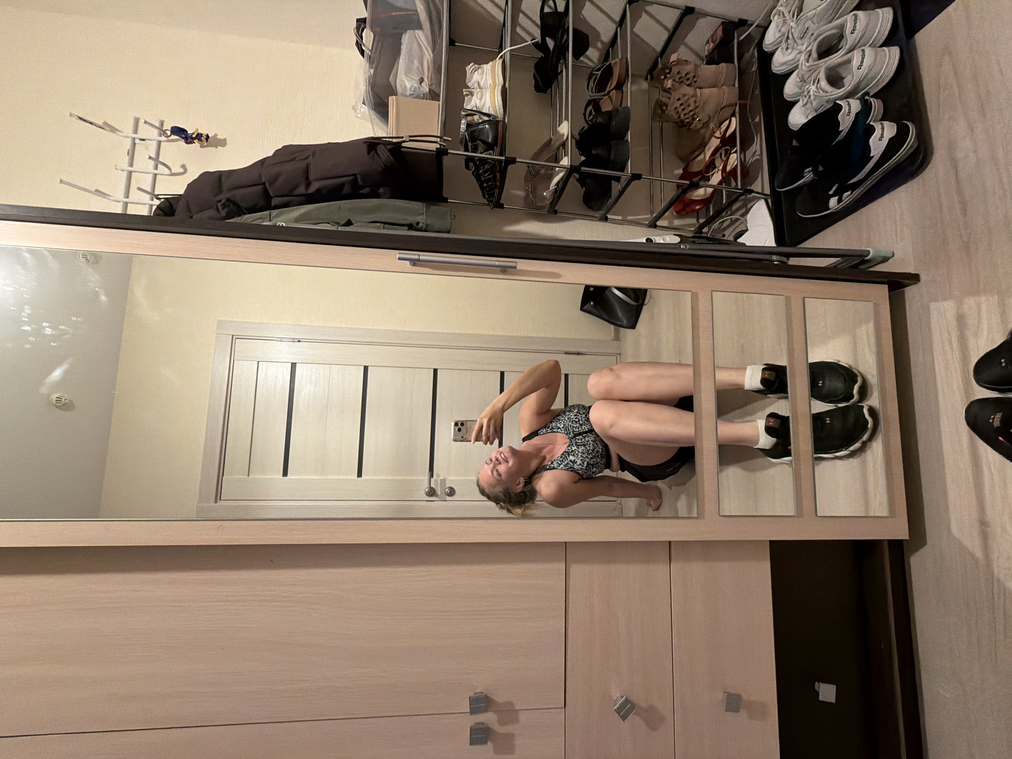 An indoor scene in Izhevsk, Russia, depicts a female subject taking a selfie in a tall, framed mirror. The subject, a woman appearing to be in her 20s or 30s with blonde hair, is seated on the floor, wearing a patterned sports bra and dark shorts. She is holding a light-colored smartphone and smiling at her reflection.
To the left of the mirror is a light-toned wooden wardrobe or cabinet with two visible sets of square, silver-colored handles. To the right of the mirror, a cream-colored wall features a white coat rack holding a dark puffer jacket and a set of keys with a yellow charm. Below the coat rack, a multi-tiered metal shoe rack is present, storing various types of footwear including sandals, heels, and athletic shoes. Above the shoe rack, several storage bins are visible.
On the floor in front of the shoe rack, a dark mat holds three pairs of athletic shoes, two white and one black/white, all appearing to be Reebok brand. A black bag is positioned on the floor near the bottom left of the mirror. The mirror itself is divided into three horizontal panels in its lower section. The floor throughout the visible area is a light wood laminate or tile. A light-colored door with dark horizontal accents and a silver handle is reflected in the mirror behind the woman. The reflected ceiling shows a white circular fixture, likely a smoke detector.