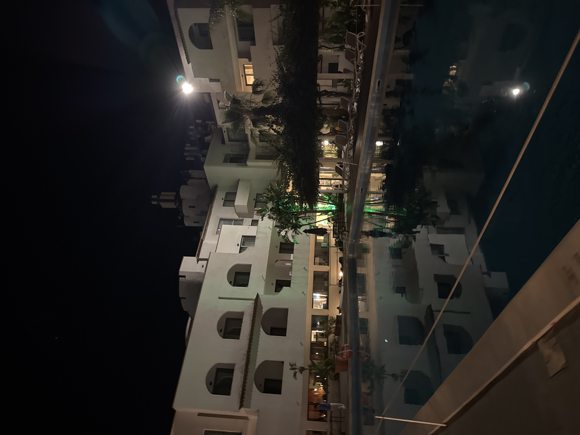The image depicts a nighttime scene at a resort in Isola delle Femmine, Italy. In the foreground, a large, dark-water swimming pool reflects the surrounding illuminated structures and artificial lights. A light-colored coping surrounds the pool, and a white rope barrier is visible across a section of the water.
Dominating the midground and background are several multi-story buildings, primarily white or off-white, featuring arched window and balcony openings, some with internal illumination. Architectural elements include a prominent tower-like structure on a building in the upper-center, and visible terra cotta roof tiles. Artificial lighting, including bright floodlights on the buildings, illuminates the complex and causes lens flares in the upper right portion of the image. Some vegetation, particularly trees, is highlighted with green accent lighting.
Numerous trees and plants, including several tall palm trees, are scattered throughout the scene. A long, dark pergola covered in dense foliage extends along the right side of the pool area, beneath which multiple light-colored lounge chairs are uniformly arranged. A dark patio umbrella and a table are also present. On the left side of the pool, a potted plant and a red and white striped lifebuoy attached to a pole are visible. One individual, wearing a light-colored top and dark trousers, is standing near the potted plant on the left. The sky above is dark and clear.