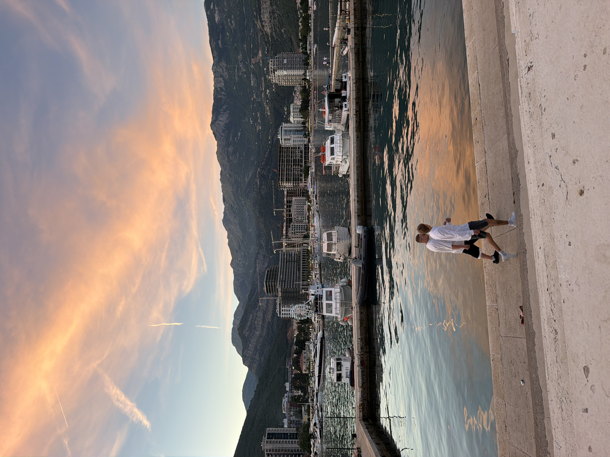In the city of Budva, Montenegro, during the golden hour, the sky displays a vibrant palette of pink, orange, and blue hues, with thin clouds and contrails from aircraft streaking across the expanse. Below, a marina is filled with various boats, including several white motorboats docked along a concrete pier. In the background, a dense cityscape with modern buildings and construction cranes rises, backed by lush, green mountains. Two men, dressed in white t-shirts and shorts, are walking along the waterfront promenade, their reflections shimmering in the calm, blue water. The foreground features a textured concrete pathway and steps.