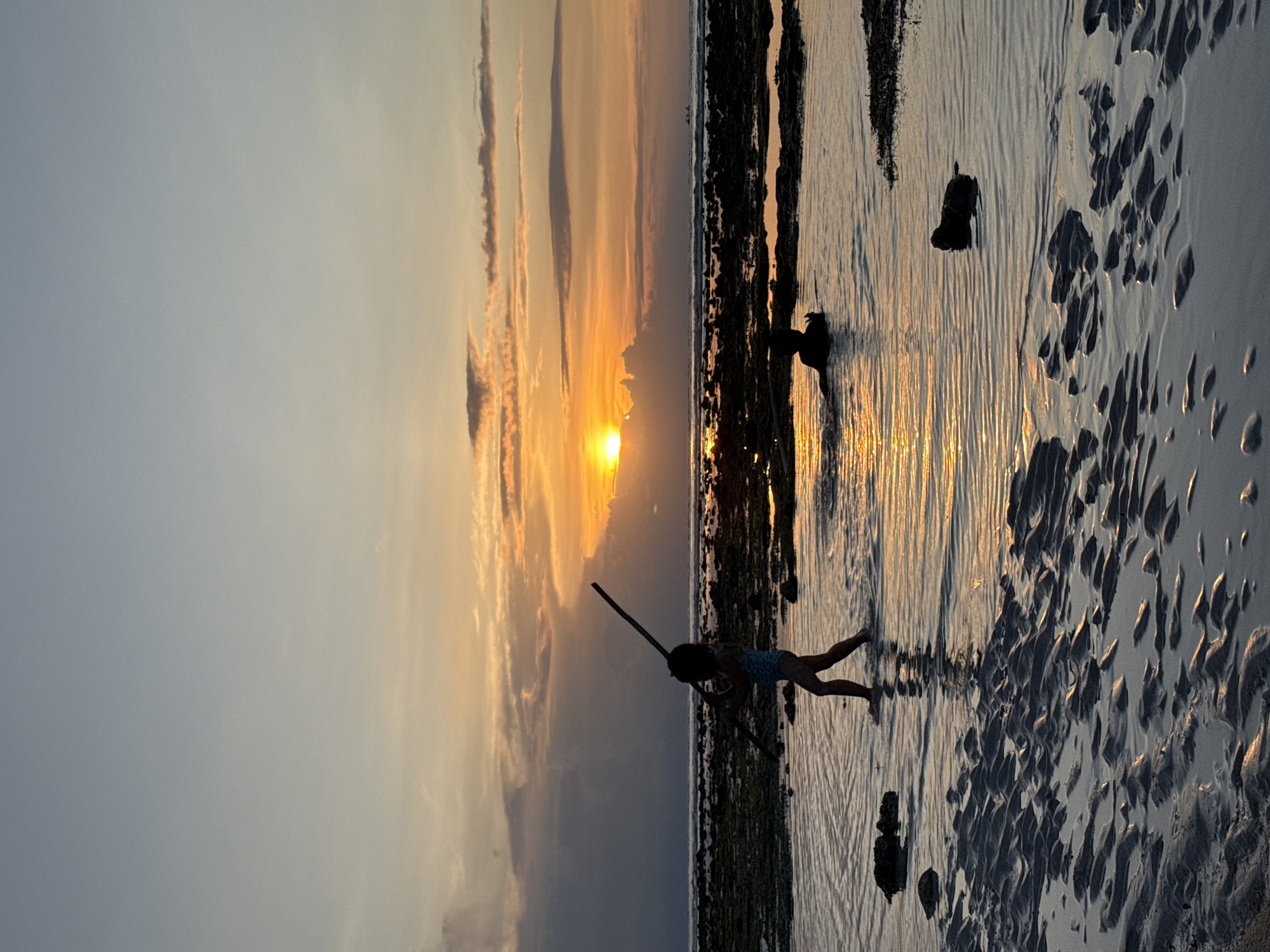 A vertical shot depicts a silhouette of a child standing on a sandy beach with wet sand formations and tidal pools reflecting the sunset. The sky features an orange-yellow glow near the horizon where the sun is setting, transitioning to lighter shades of yellow and blue higher up. Several long, thin clouds streak across the sky, parallel to the horizon. The silhouette of the child holds a long stick. The location is Unknown, Indonesia.