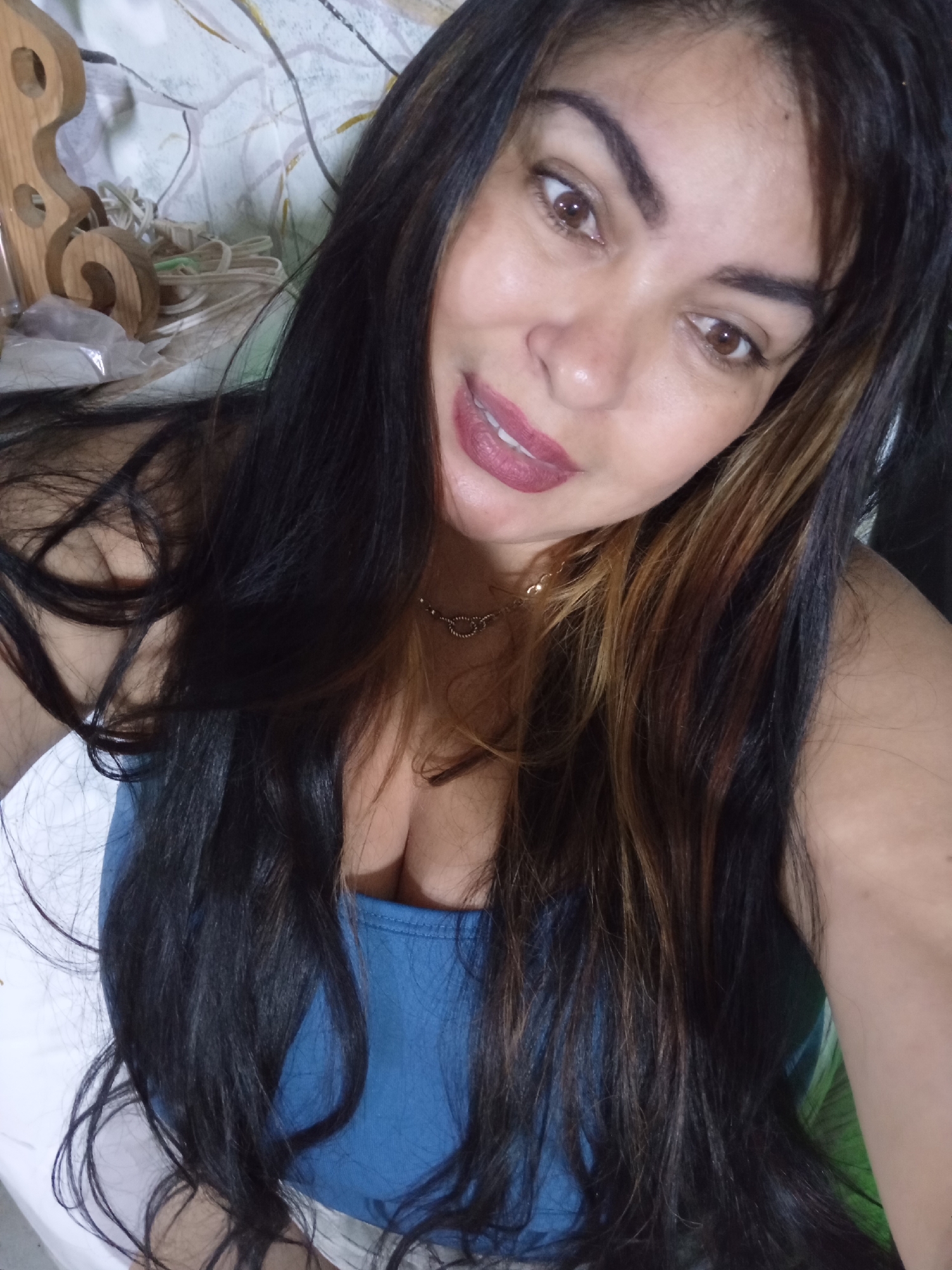 This image captures an adult female in an indoor setting within Juan Griego, Venezuela. The subject is positioned for a chest-up portrait, looking directly at the viewer. She has long, dark hair with streaks of brown, worn down and flowing over her shoulders and chest. Her features include brown eyes, defined eyebrows, and lips adorned with dark pink lipstick. Her mouth is slightly open with the tongue partially visible, conveying a subtle smirk. She is attired in a blue scoop-neck top, revealing cleavage, and a gold-toned necklace with small heart-shaped links. The background displays a light-colored wall featuring a faint, abstract botanical pattern. To the upper left, a carved wooden decorative object with curved edges and apertures is visible. Below this, a cluster of white cables or wires is present. The scene is evenly lit, indicative of indoor lighting.