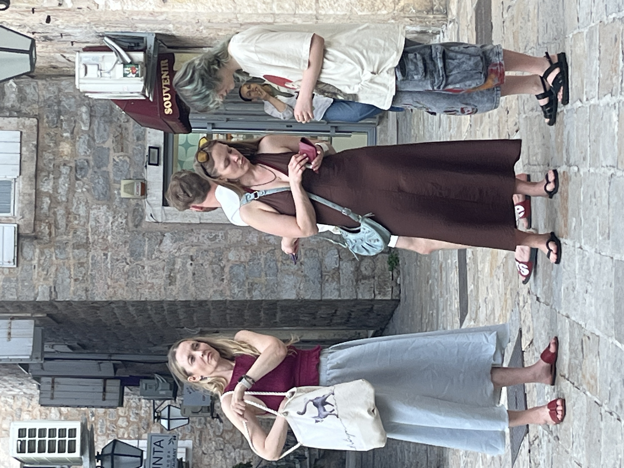 In Budva, Montenegro, a group of people are gathered on a cobblestone street lined with stone buildings. A woman on the left, wearing a maroon top and a light blue skirt, holds a beige canvas tote bag with a cat illustration. To her right, another woman in a brown dress is looking at her phone. A third person with blue-tinted hair and a light-colored t-shirt stands to the right of the woman with the brown dress. In the background, a woman is visible smoking outside a shop with a "SOUVENIR" awning. The street appears to be a pedestrian area with decorative lighting and signage.