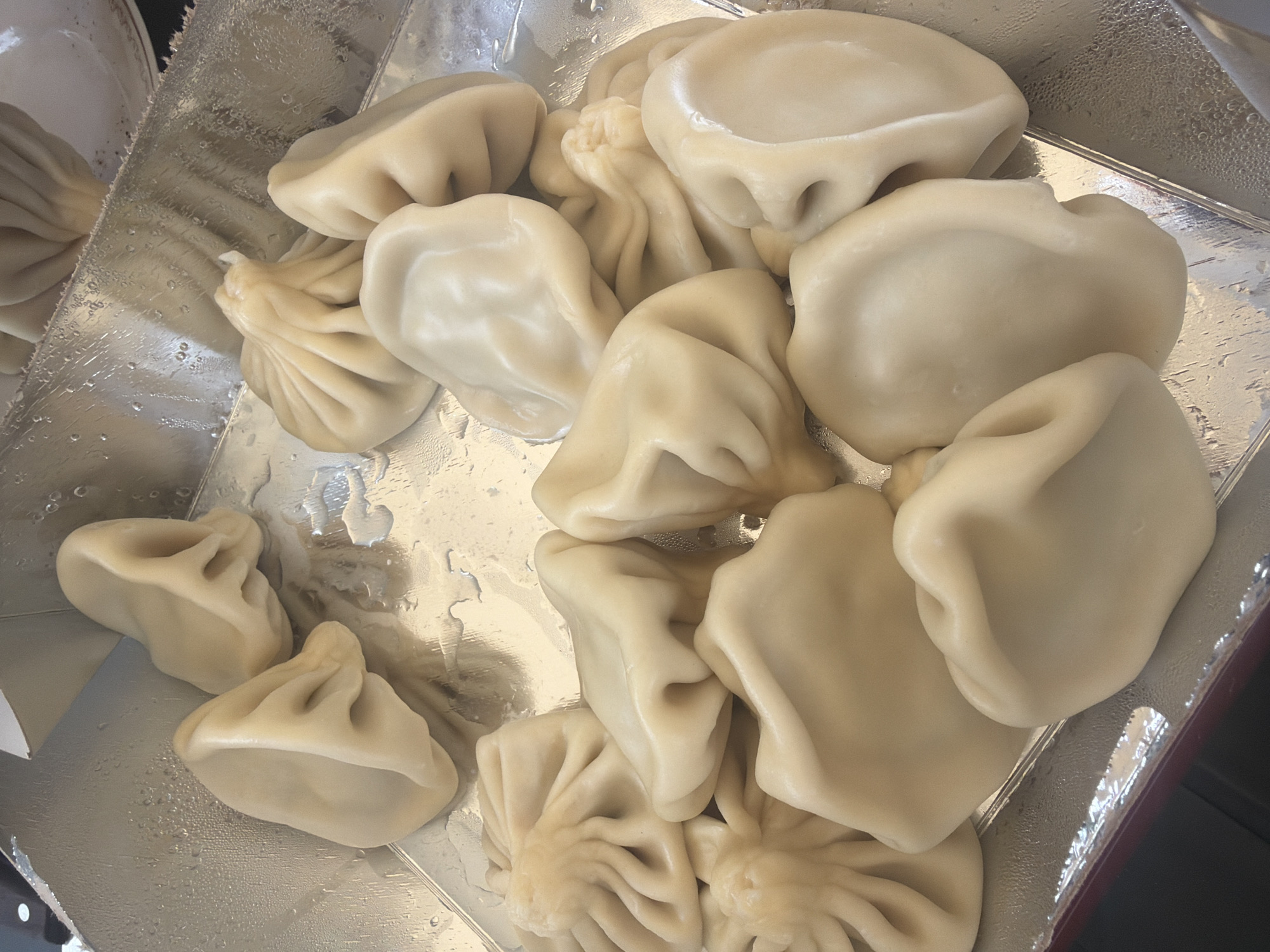 A close-up, high-angle shot depicts a collection of boiled khinkali (Georgian dumplings) arranged in an aluminum container, possibly for transport or serving. The khinkali vary in size and shape, with some displaying the characteristic pleated tops and others appearing smoother and more filled out. The dumplings are pale in color, suggesting they are freshly cooked. Small droplets of water or condensation are visible on the reflective surface of the container. The overall composition suggests a food service setting, potentially in Dusheti, Georgia, a location known for its khinkali.