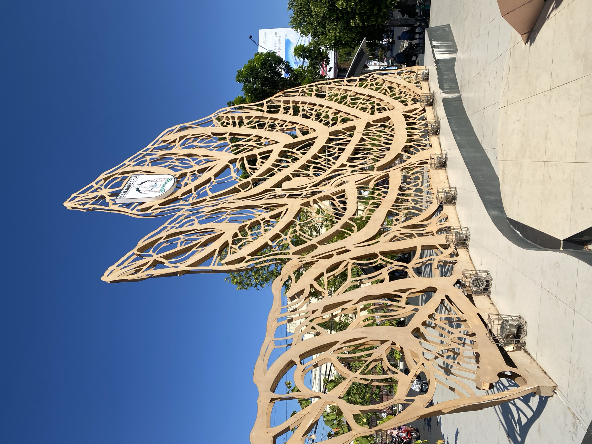 The image shows a large, abstract sculpture with intricate, branch-like patterns, made of a light brown material. The sculpture is located outdoors in a public space with a bright blue sky overhead. A circular emblem with the text "BANJARBARU" is prominently displayed on the upper part of the sculpture. In the background, there are trees, buildings, a billboard, and several people and vehicles on a street. The foreground consists of a paved area with tiled surfaces and a raised concrete platform. Several small, caged lights are positioned along the base of the sculpture. The overall scene suggests a sunny day in the city of Banjarbaru, Indonesia, at a public monument or park.