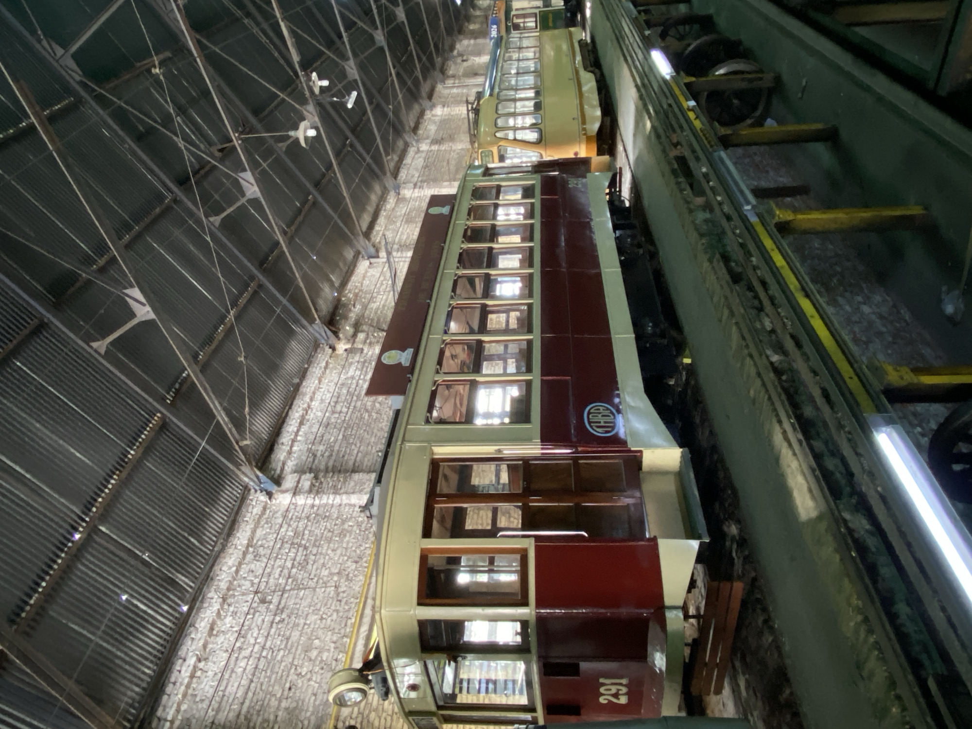 The image depicts the interior of a tram depot in Buenos Aires, Argentina. Several vintage trams are visible, the closest one numbered "291" with a livery of light green and maroon. The depot has a corrugated metal roof supported by a framework of steel beams. The ground appears to be paved with stone or brick. Some of the trams are parked over a maintenance pit along a dark green and yellow rail system. Bright fluorescent lights are installed along the side walls. The scene is dimly lit.