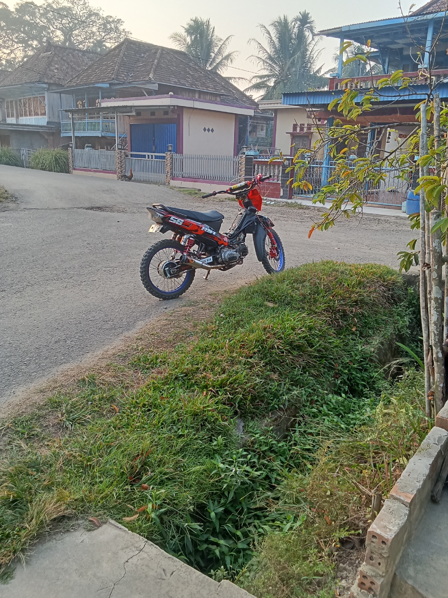 The image depicts a rural roadside scene in Tebing Bulang, Indonesia, with a modified red and black dirt bike parked on the left side of a paved road. The motorbike is customized with off-road tires and an aftermarket exhaust. The background shows residential buildings with tiled roofs and fences. Lush green vegetation, including grass and trees, lines the roadside and slopes down into a ditch. The sky is clear, suggesting it is daytime. No people are visible in the image.