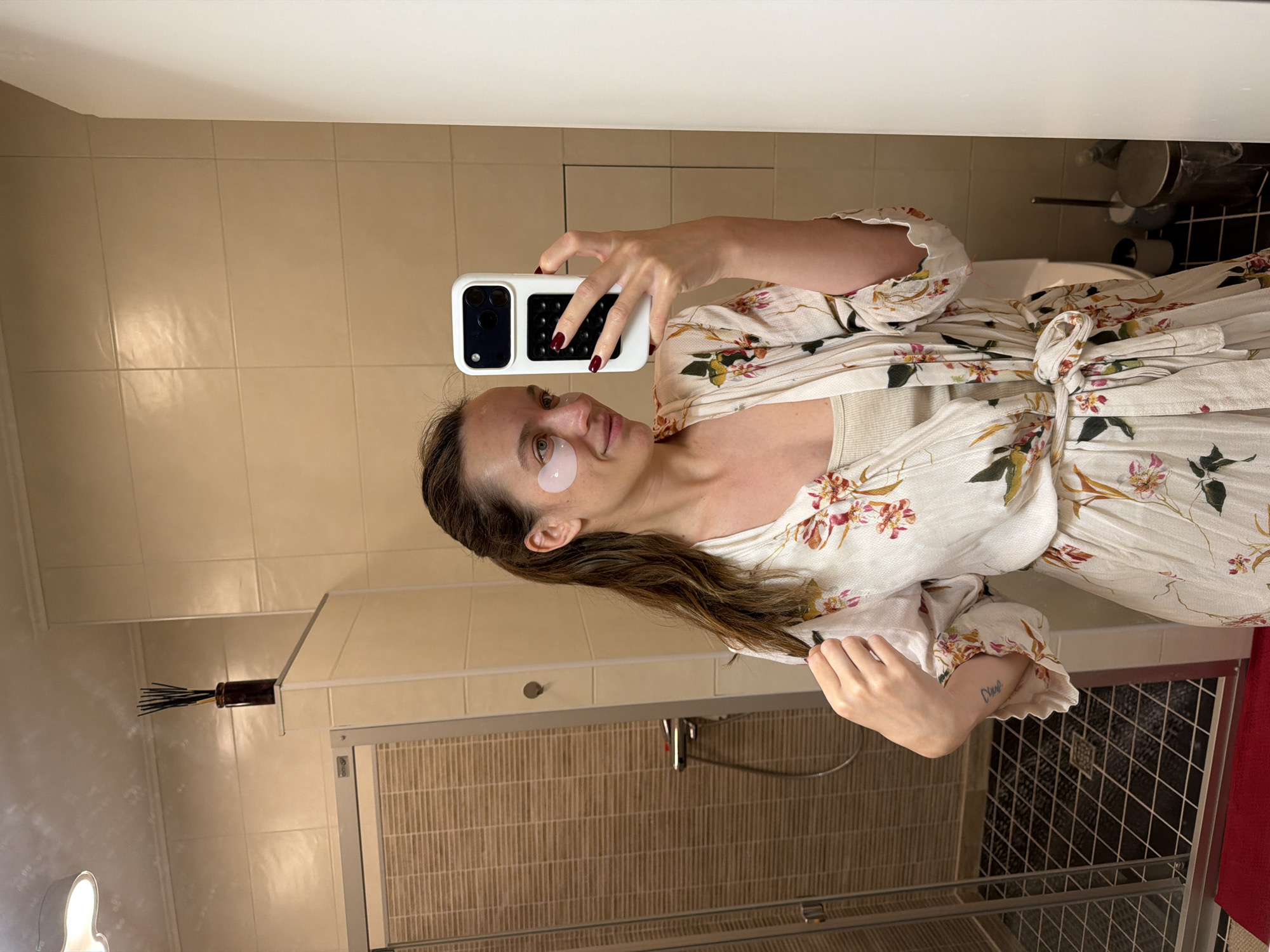A woman stands in a bathroom and takes a selfie. She wears a floral robe over a top, and an eye patch on her left eye. She is holding a white smartphone. The bathroom has beige tiles, a shower with a sliding door, and a red curtain visible. A dark, cylindrical object with sticks is mounted on the wall. There is no evidence to suggest that the scene is located in Tarasovka, Russia.