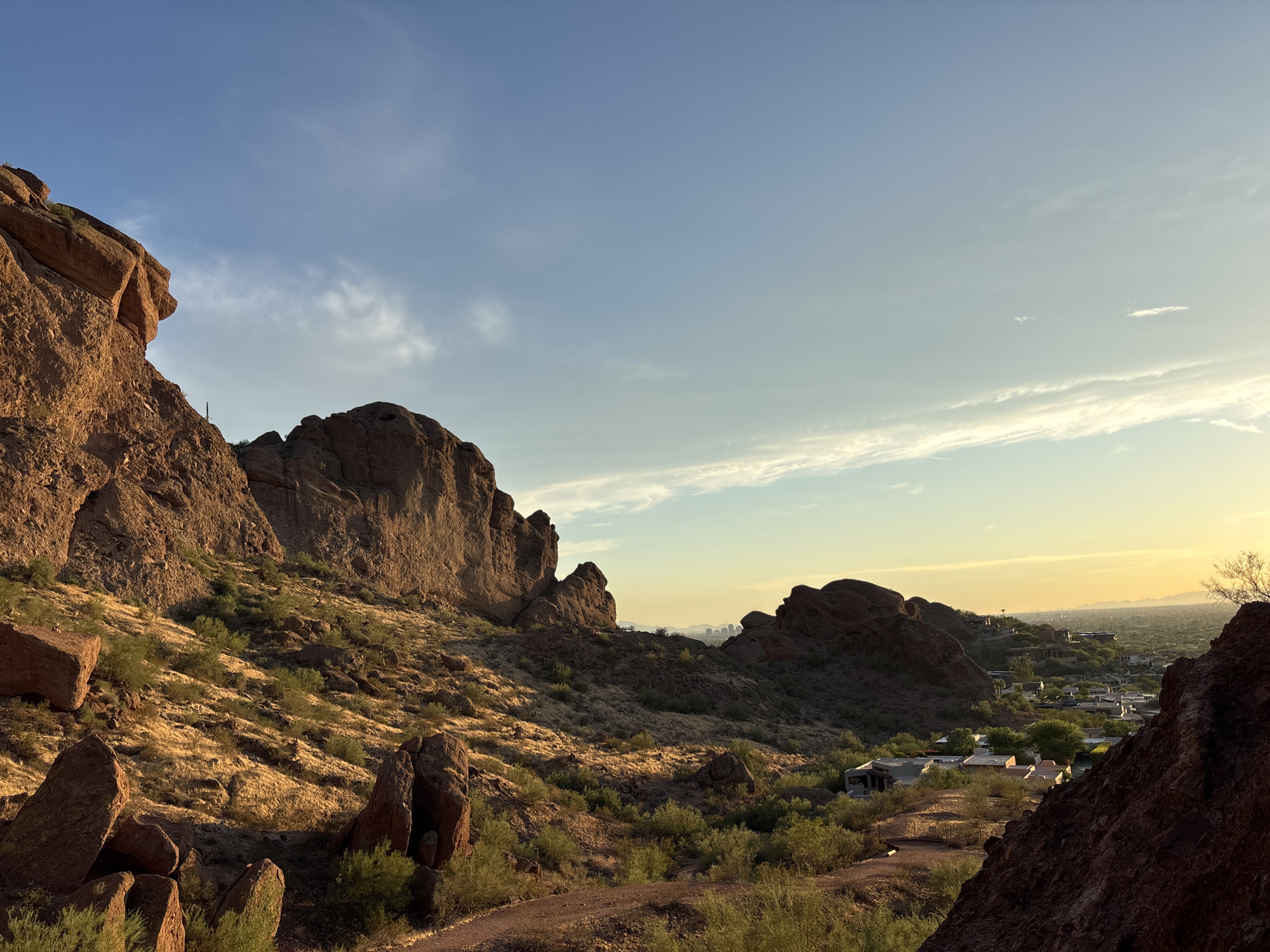 The image depicts a scenic desert landscape at golden hour, likely in the South Mountain Park and Preserve in Phoenix, Arizona. Towering red rock formations dominate the left and foreground of the image, casting long shadows. Sparse desert vegetation, including scrub brush and dry grasses, covers the undulating terrain. A dirt path winds through the scene in the foreground. In the distance, nestled in the valley below, a suburban development with houses and trees is visible, and beyond that, the faint silhouette of the Phoenix skyline can be seen against the warm, hazy horizon. The sky is a gradient of blue, with wispy clouds catching the last rays of the setting sun, which casts a golden glow over the entire landscape. There are no people or animals visible in the image.