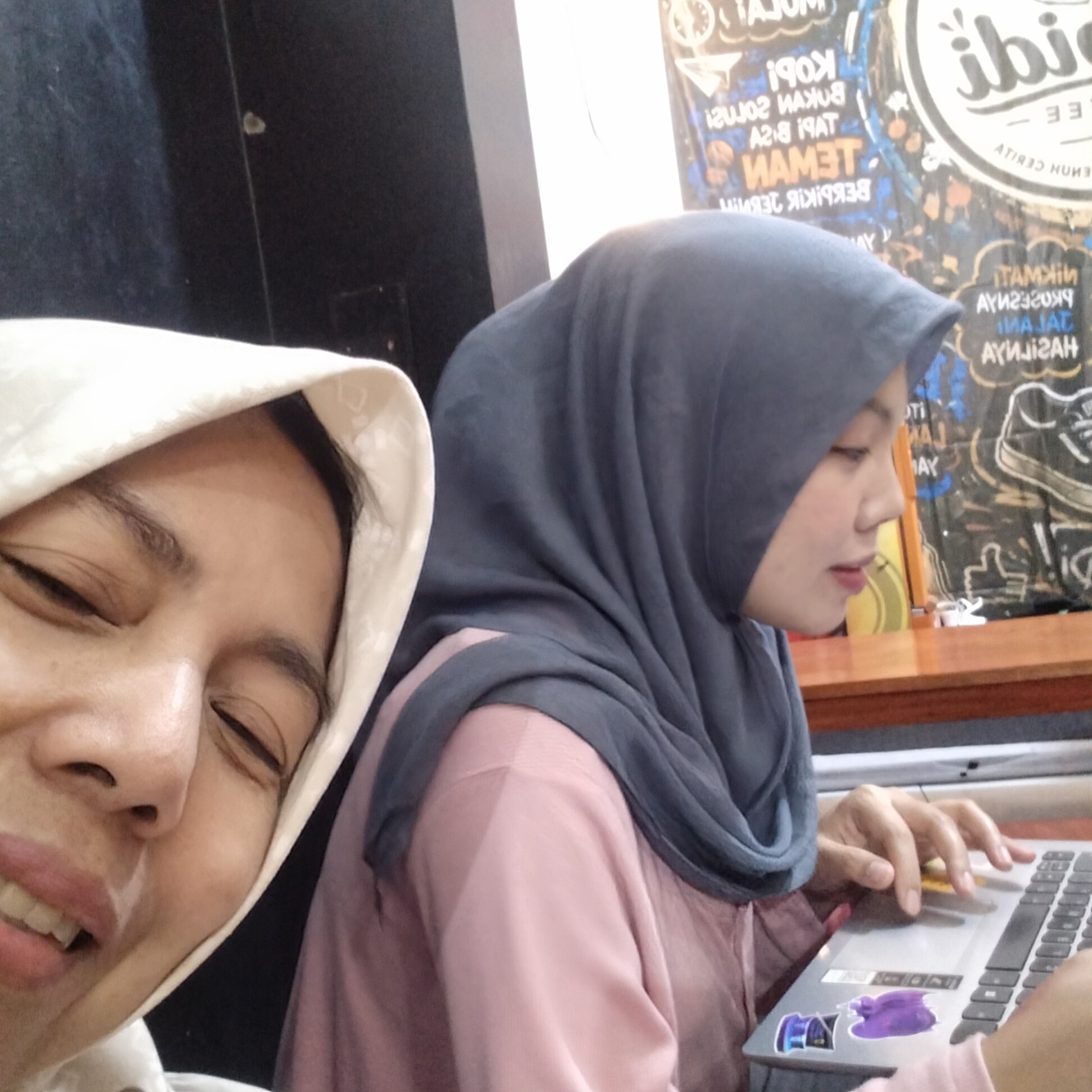 Two women are in a room, possibly a cafe, with a poster on the wall behind them. The woman in the foreground is on the left, wearing a light-colored hijab. She is looking downwards and to her right, with her eyes closed and her mouth slightly open, revealing her teeth. Her skin has a light complexion and appears to have some freckles. The woman in the background is on the right, wearing a dark blue hijab and a pink shirt. She is looking down and to her left, focusing on a silver laptop that she is using with her right hand. The laptop has several stickers on it. The background poster is colorful and appears to have text in Indonesian, some of which is upside down or reversed. The furniture visible includes a wooden table in front of the woman using the laptop. The lighting suggests an indoor setting. The specific location is not identifiable from the image itself.