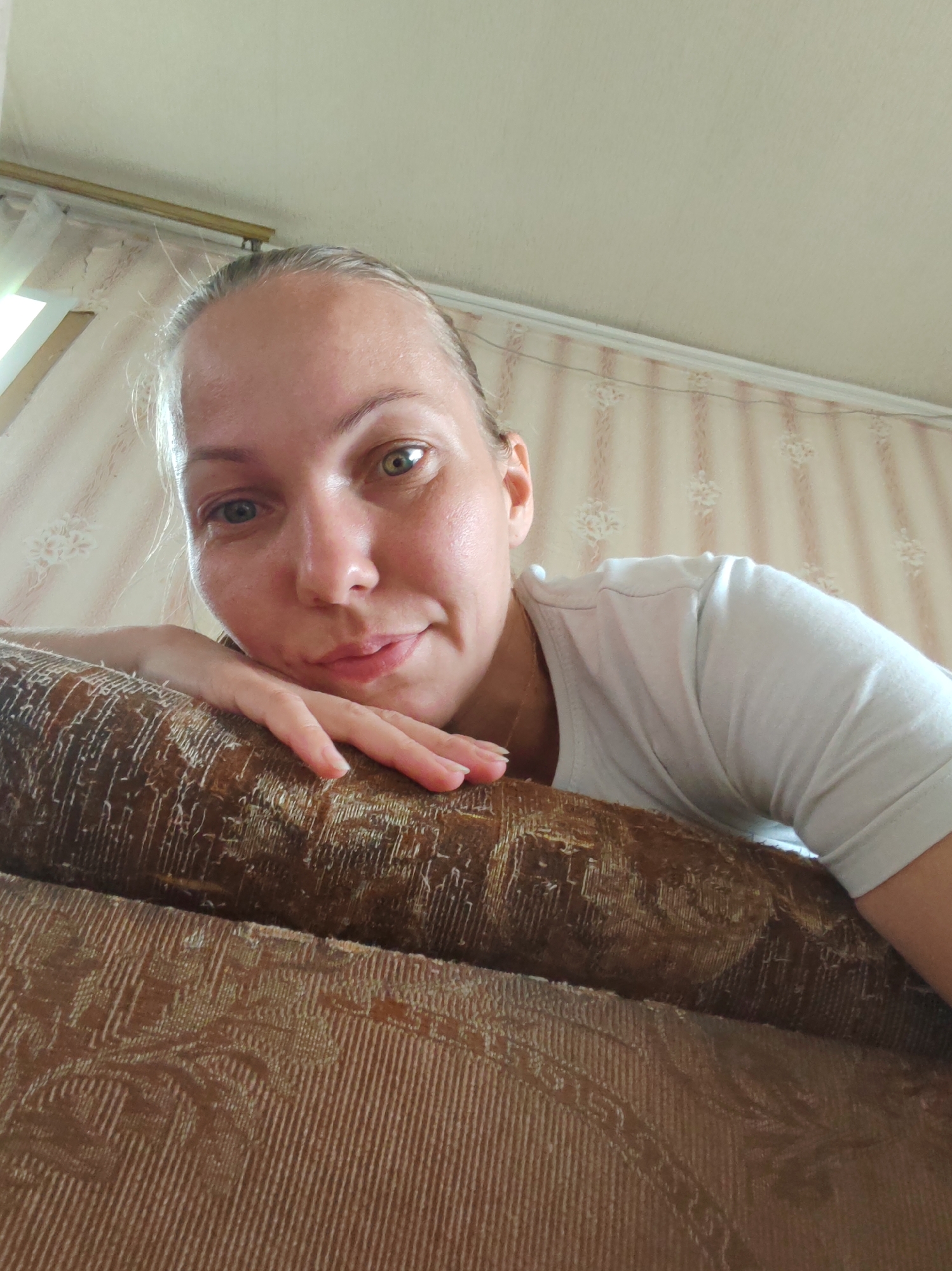 The image is a selfie taken from a low angle, looking up at a woman lying on her side. She is resting her head on her arm, which is draped over a textured, brown sofa. The woman has light-colored hair pulled back, and she is wearing a white t-shirt. Her face is partially obscured by her arm, but her eyes are visible, with heterochromia (one eye is lighter than the other). She is looking towards the camera with a slight smile. The background consists of a wall with striped wallpaper and a window with sheer curtains. The overall impression is one of casual domesticity. The location is specified as Чухлэм, Russia.