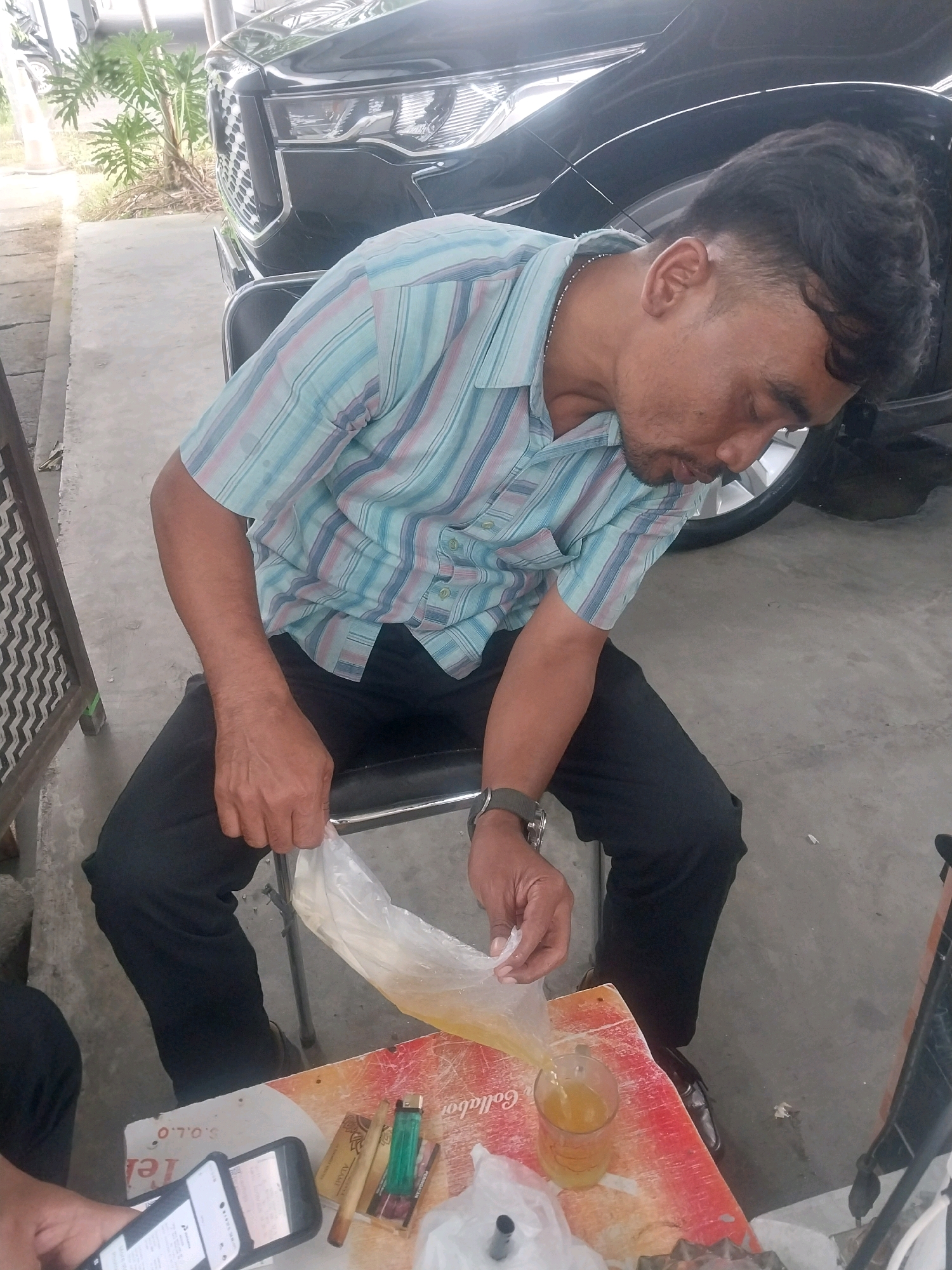 A man in a striped collared shirt and black pants is seated on a chair. He is holding a clear plastic bag filled with a yellow liquid and pouring it into a glass on a table. A phone, cigarettes, a lighter, and a watch are also on the table. In the background, a black car is parked. The scene appears to be an outdoor setting with a concrete floor and some vegetation visible. The overall impression is that of a casual scene in Sleman, Indonesia.