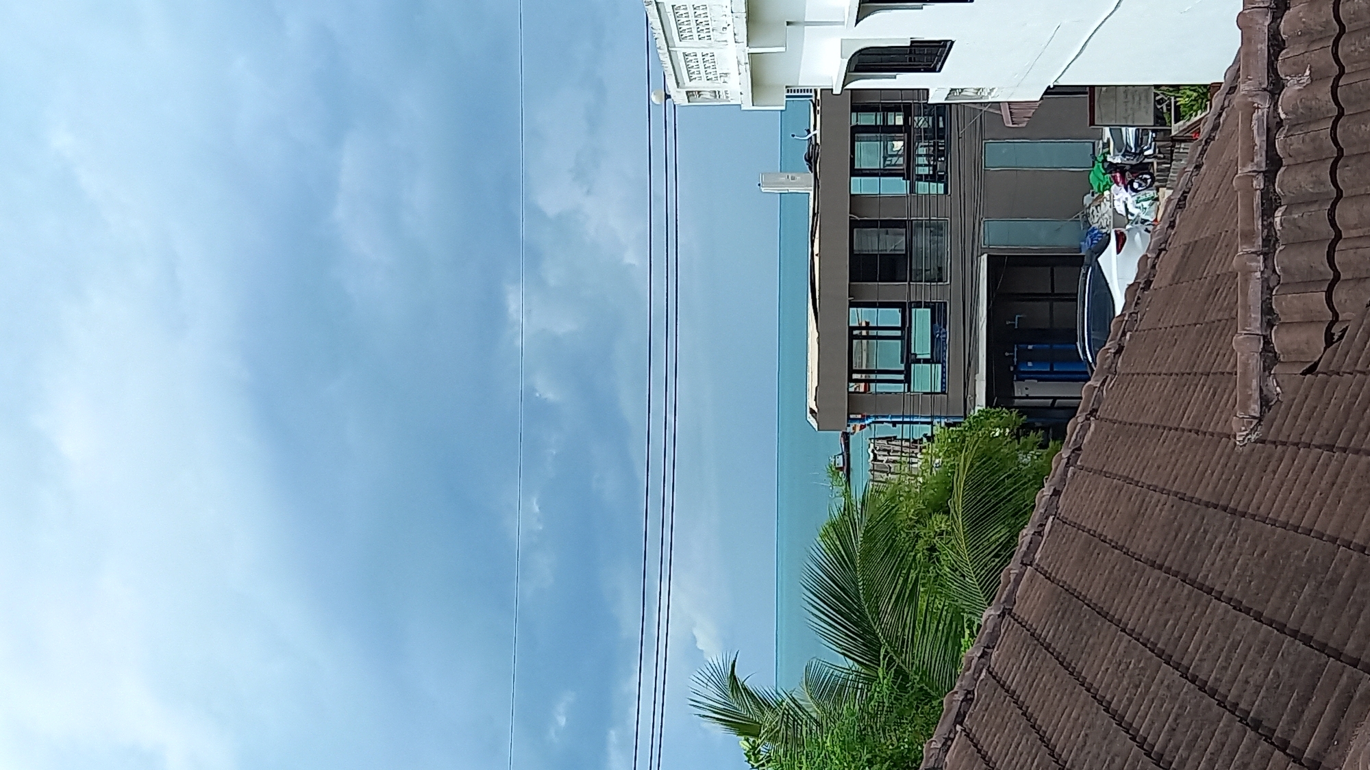 A cloudy day over a residential area on Ko Pha-ngan, Thailand. The scene is dominated by a tiled brown roof in the foreground, with a modern, multi-story building with large windows and a white building with decorative railings visible behind it. Beyond these structures, a body of turquoise water, likely the sea, stretches to the horizon under a heavy, gray-blue sky with scattered clouds. Palm fronds from a lush green tree emerge from the left side of the frame. A white car and motorcycles are parked near the buildings.