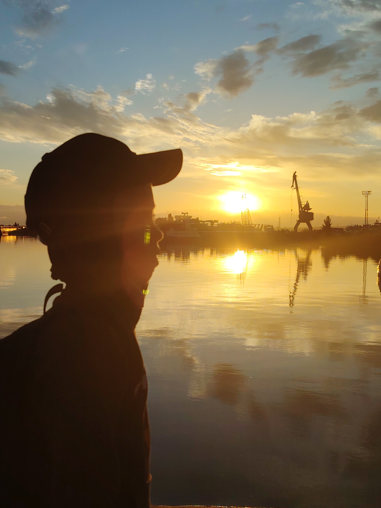 This is a medium shot, taken from the right, of a person wearing a dark baseball cap and jacket, silhouetted against a bright sunset over a body of water. The person is looking out towards the right of the frame. The sun is low on the horizon, casting a golden glow across the sky and reflecting vibrantly on the water. In the background, on the far shore, a crane and various industrial structures are visible, suggesting a port or industrial area. The sky is partially filled with scattered clouds, some of which are illuminated by the setting sun. The water is calm, with a clear reflection of the sky and the structures on the shore. The overall scene captures a moment of quiet contemplation at dusk in an industrial waterfront setting, likely in Chaikovsky, Russia, given its known port facilities on the Kama River.