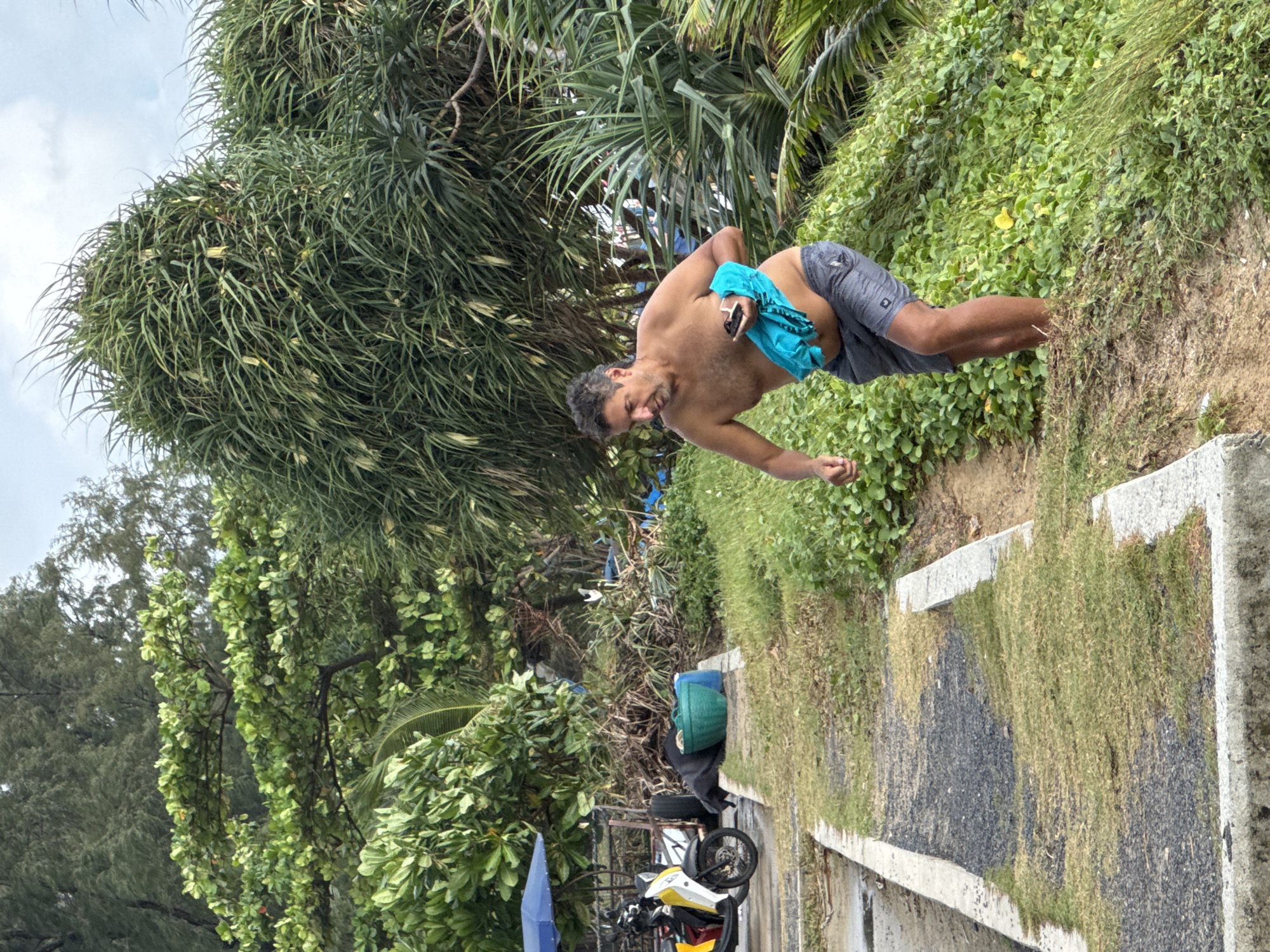 A man is walking down a grassy slope in Karon, Thailand. He is shirtless and wearing grey swim trunks, and he is holding a folded blue cloth and a black object in his hands. The man appears to be mid-stride, with his left leg forward. The background consists of dense green foliage and trees. To the left of the man, a paved path with overgrown grass runs alongside a body of water. A parked scooter with a blue umbrella is visible in the distance. The sky is overcast.