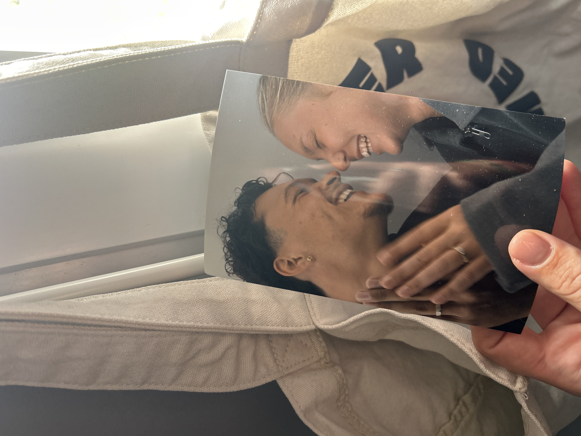 A photograph depicting a smiling mixed-race couple is being held over an open cream-colored canvas tote bag. The woman in the photo has short blonde hair, and the man has short dark curly hair. Both are wearing rings. The bag has black lettering on it. The location, Lannoy, France, is not discernible from the given image.