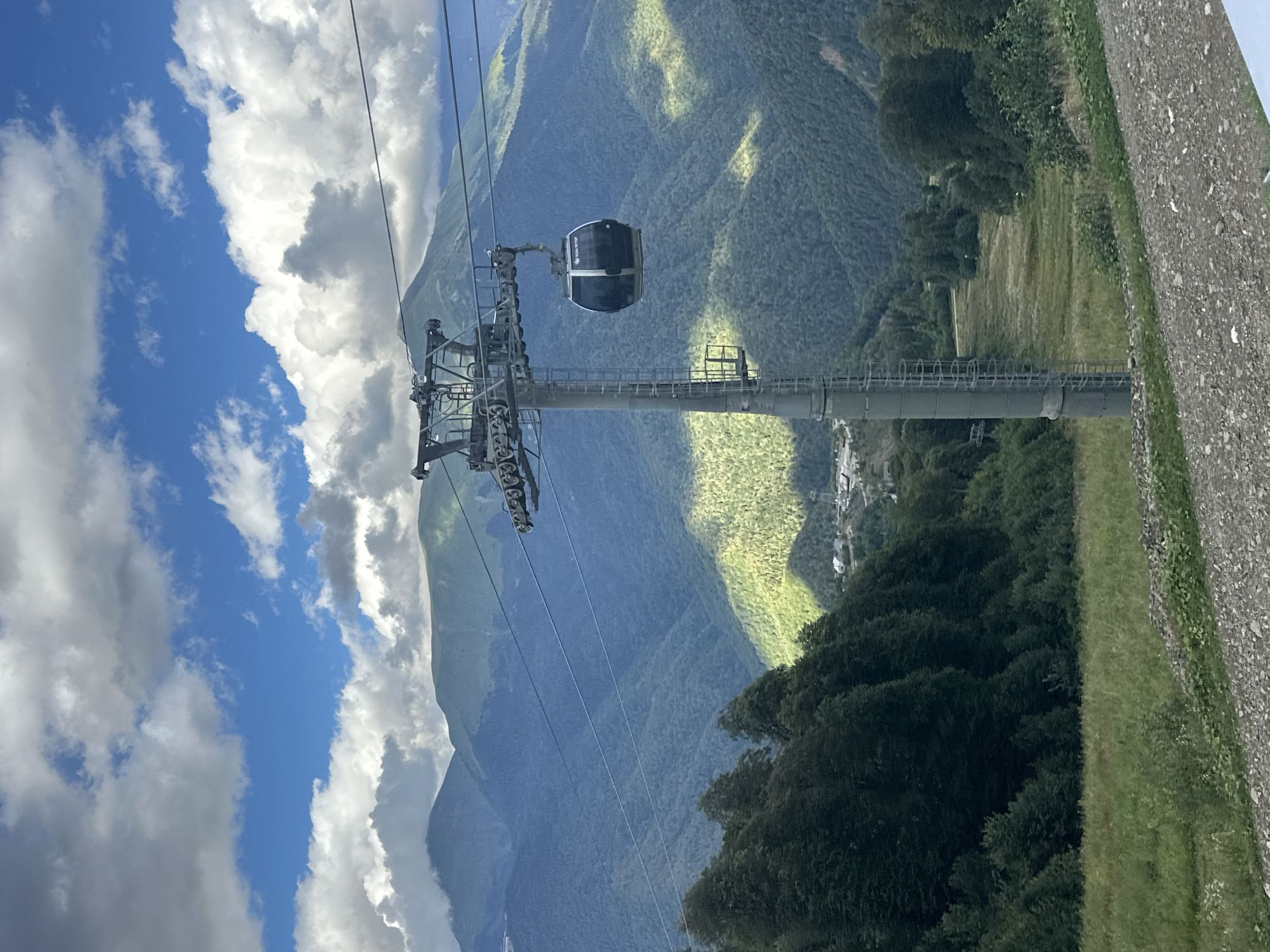 The image captures a scenic view of the Rosa Khutor ski resort in Estosadok, Russia. A modern cable car cabin is suspended from a steel tower, traversing over a lush, green mountainous landscape. The sky is adorned with a mix of fluffy white and grey clouds against a clear blue backdrop. The mountains, densely covered with evergreen trees, recede into the distance, with sunlight casting dappled shadows on their slopes. In the foreground, a gravel path leads the eye towards the mountainous terrain, hinting at the recreational activities available in the area. The scene is one of natural beauty enhanced by the presence of ski resort infrastructure.