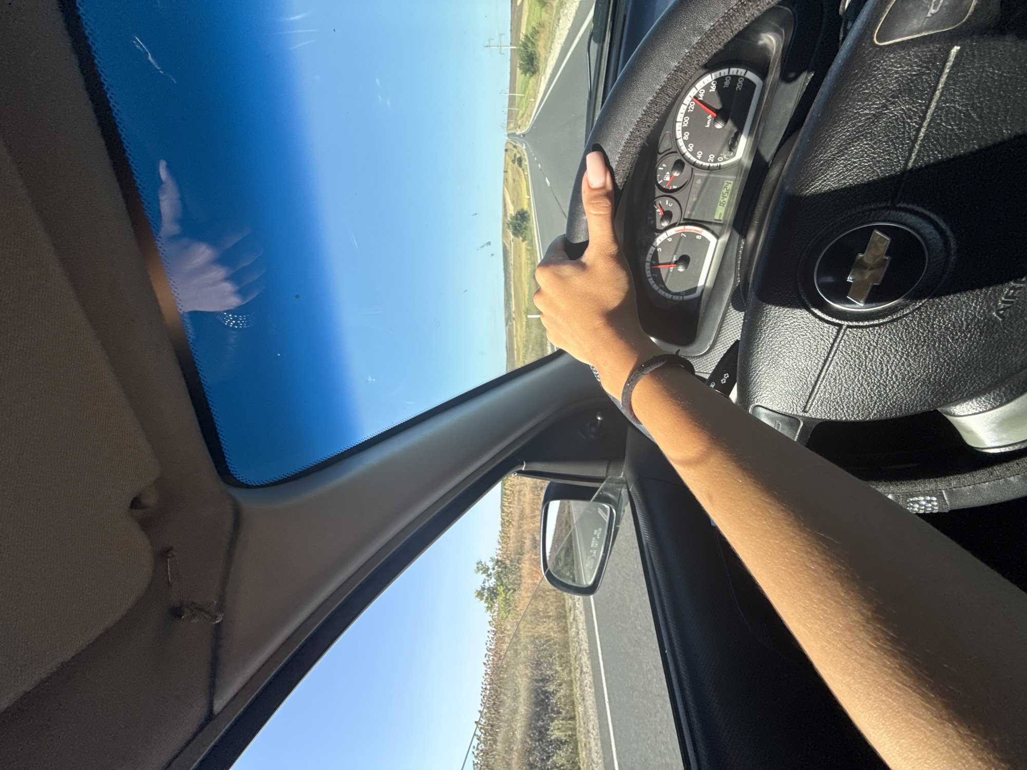 A first-person perspective shows a person driving a Chevrolet vehicle on a clear, sunny day. The driver's right hand, adorned with light pink nail polish and a dark bracelet, rests on the steering wheel. The car's dashboard displays a speedometer indicating approximately 120 km/h, an odometer reading 164524, and a tachometer. The view through the windshield reveals a straight, paved road stretching into the distance, flanked by dry, yellowish fields and scattered trees. Power lines are visible along the side of the road. The car's interior includes a side mirror and a portion of the headliner with a safety pin attached. The overall scene suggests travel on a rural road, possibly near Comrat, Moldova, given the characteristic landscape of the region.