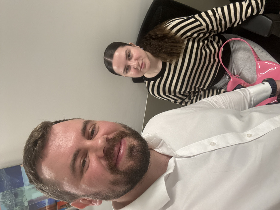The image displays two individuals, one male and one female, in an indoor setting in Dubai, United Arab Emirates. The male, positioned in the foreground, has a beard and dark hair, and wears a white collared shirt with the left cuff featuring "1983" embroidery. He holds the camera, appearing to take a selfie. The female, seated behind and to his right, has dark hair styled in a crimped low ponytail. She wears a black and off-white horizontally striped long-sleeved sweater and light-colored jeans. A glossy, bright pink shoulder bag rests on her lap. She is seated in a dark office chair. The background features a plain light-colored wall with a partial view of an abstract artwork in the upper left, incorporating blue, orange, and white elements.