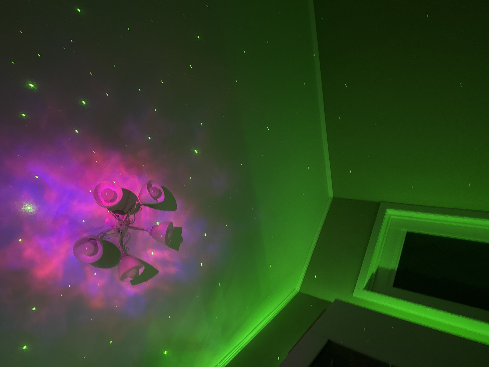 A low-angle shot depicts a room with a chandelier projecting a violet and red nebula pattern onto the ceiling, which is also speckled with small green lights. The walls are green. A window frame is visible in the corner of the room. The location appears to be indoors and artificially illuminated, but no people or specific location can be determined from the image.