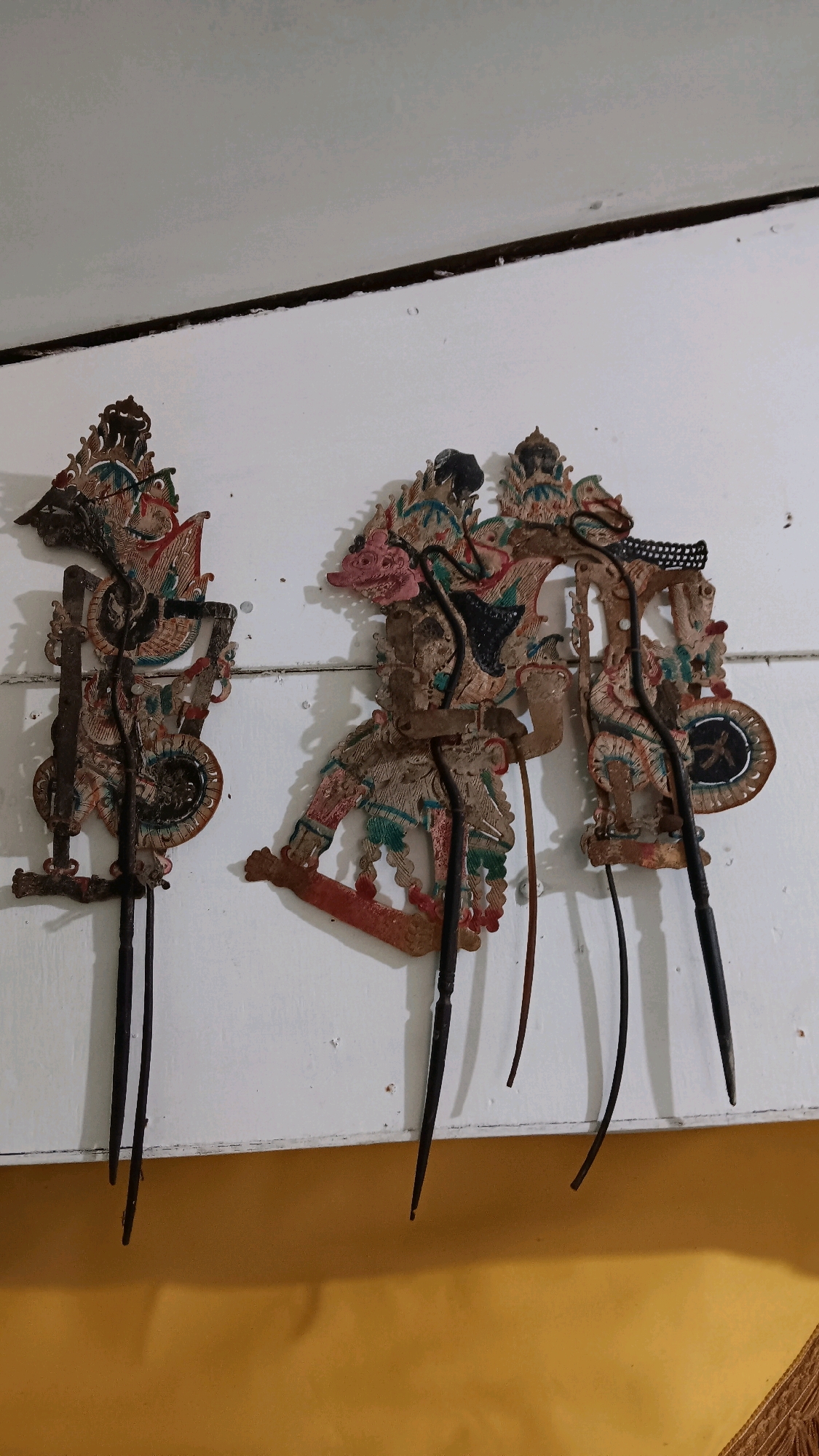 The image displays three traditional Indonesian wayang kulit puppets hanging on a white wooden wall. The puppets are intricately carved and painted, featuring detailed characters with elaborate headdresses and traditional clothing. They are mounted on slender rods for manipulation. The wall appears to be part of an interior space with a dark horizontal line running across it near the top. Below the wall is a bright yellow surface, possibly a floor or a lower wall, with a visible shadow cast by the puppets. The lighting suggests an indoor environment. The specific location within Depok, Indonesia, cannot be determined from the visual information provided in the image.