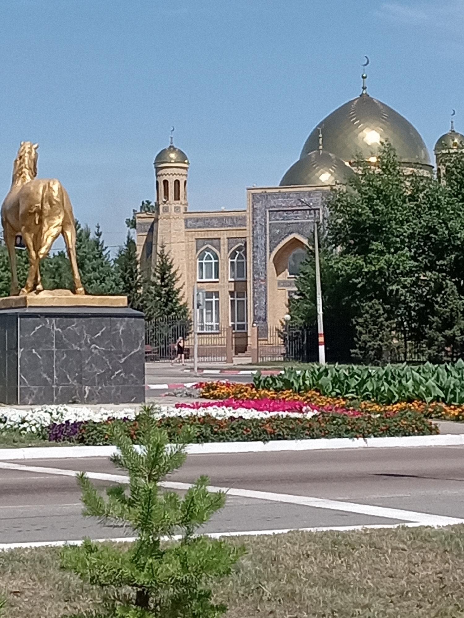 The image shows a gold-colored statue of a horse, facing away from the viewer, on a dark, marble-like pedestal. Behind the statue is a tan-colored building with a large golden dome and smaller domes, characteristic of a mosque, with minarets. The building has arched windows and decorative tilework. Trees, including evergreen trees, are visible behind the building and to the right. In the foreground, there is a flowerbed with colorful flowers, including white, pink, and yellow blooms, separated by a white border. A road with white lane markings is in front of the flowerbed. A small evergreen tree is in the lower left foreground. The sky is clear and blue. The scene is outdoors on a sunny day. This is likely the Central Mosque in Rudny, Kazakhstan, given the architectural style of the mosque and the presence of the horse statue, which is a significant monument in the city.