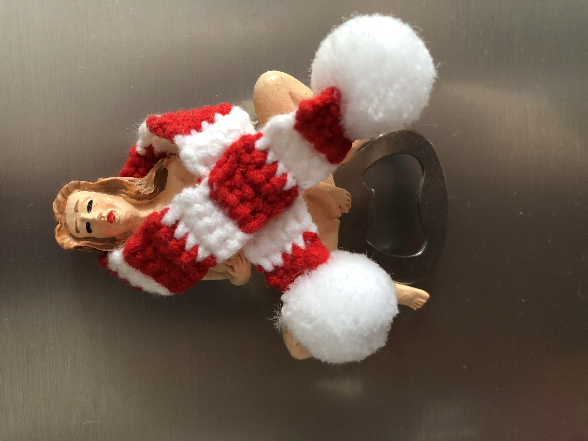A decorative bottle opener is attached to a metallic surface. The bottle opener is designed in the shape of a seated nude female figurine with tan skin and light brown hair. She is wearing a red and white striped scarf around her upper body and two large white pom-poms, one on each side of her lower body. The bottle opener mechanism is visible beneath the figurine. The background is a brushed metallic surface, likely a refrigerator door. The lighting suggests it is daytime, casting a slight shadow.