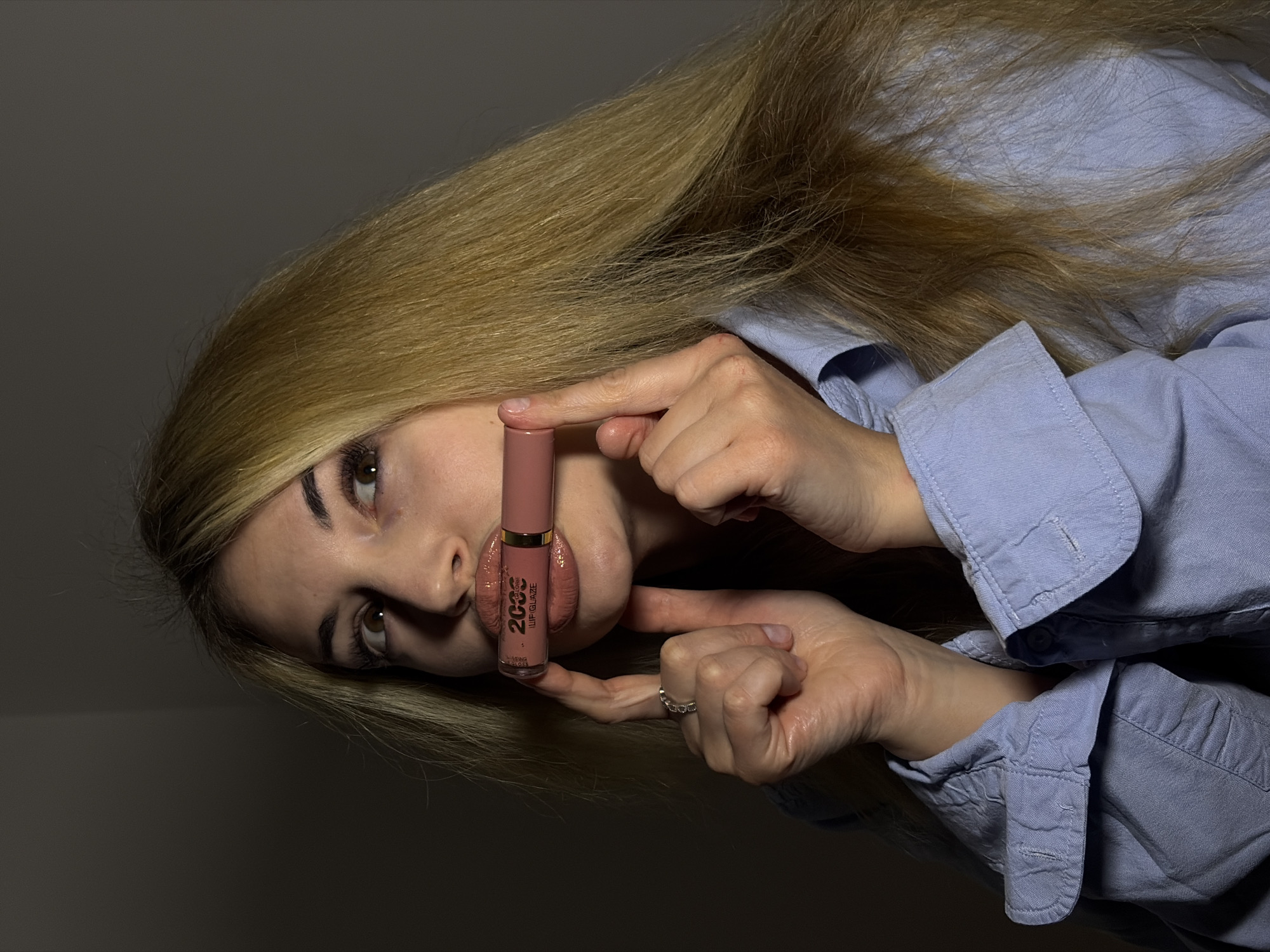 The image displays a close-up portrait of a young woman applying or posing with a lip gloss, oriented diagonally. Her face is seen in profile, with her gaze directed upwards and to the right. She has long, blonde to light brown hair that flows around her head and shoulders, and she is wearing a light blue, long-sleeved button-up shirt.
The woman's hands hold a light pink or nude-colored lip gloss applicator bottle between her glossy, slightly parted lips. A silver-toned ring is visible on the ring finger of her right hand. The text "2000" and "LIP GLAZE" are clearly visible on the side of the lip gloss bottle, with further smaller, less distinct words beneath. Her eyes show visible makeup, including eyeliner and mascara, and her eyebrows are neatly shaped.
The background is dark and featureless, indicating an indoor setting in Budva, Montenegro, likely illuminated by artificial, directional lighting that creates distinct shadows and highlights on her face, hair, and hands. The overall impression is a focused and stylish depiction of a beauty product application.