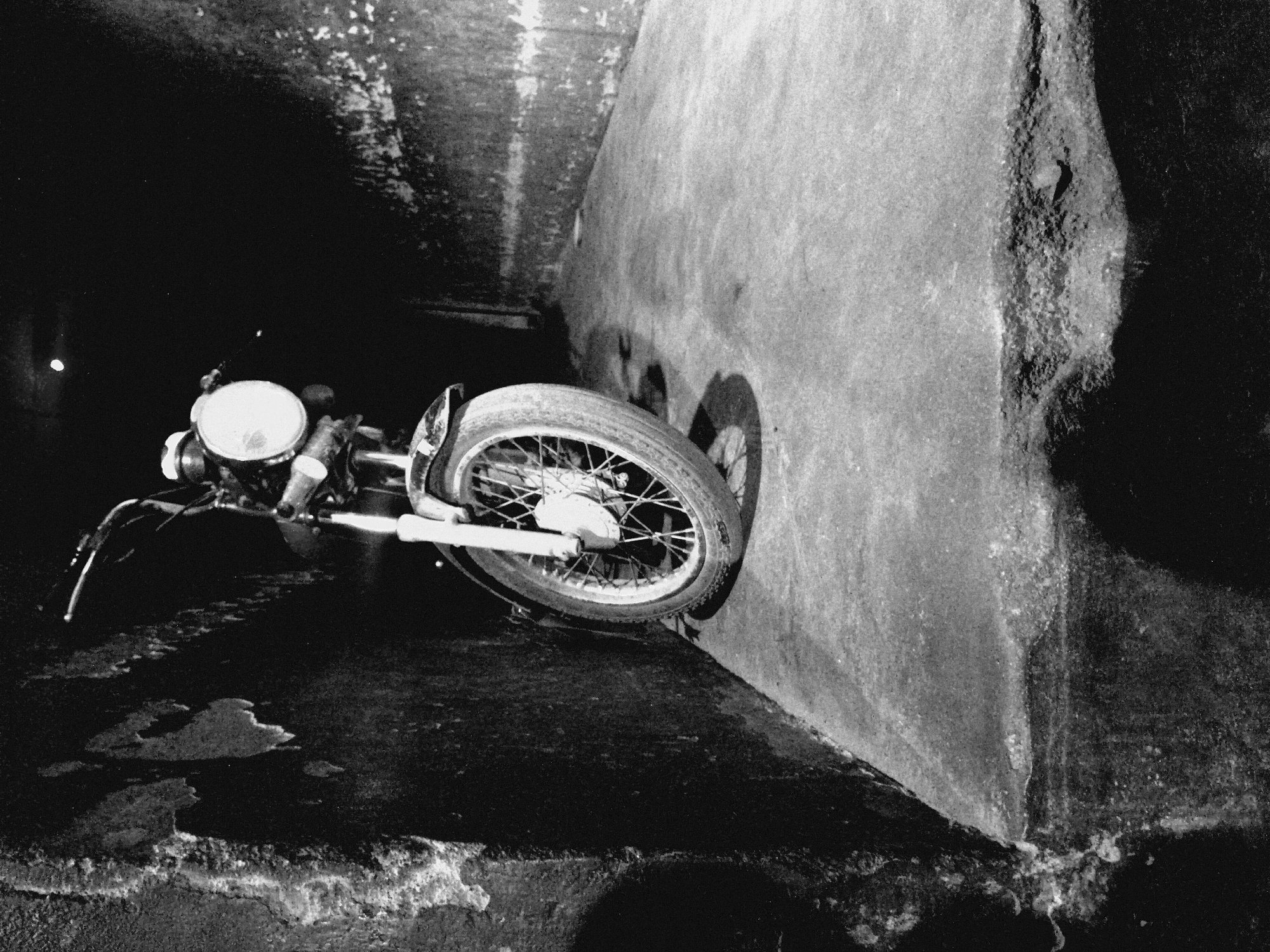 The image is a black and white photograph depicting a motorcycle crashed against a concrete wall within what appears to be a tunnel or underpass. The motorcycle is positioned at an angle, with its front wheel pressed against the wall. The environment is dimly lit, with the upper portion of the tunnel being shrouded in darkness. The ground is uneven and appears wet. The scene is devoid of people.