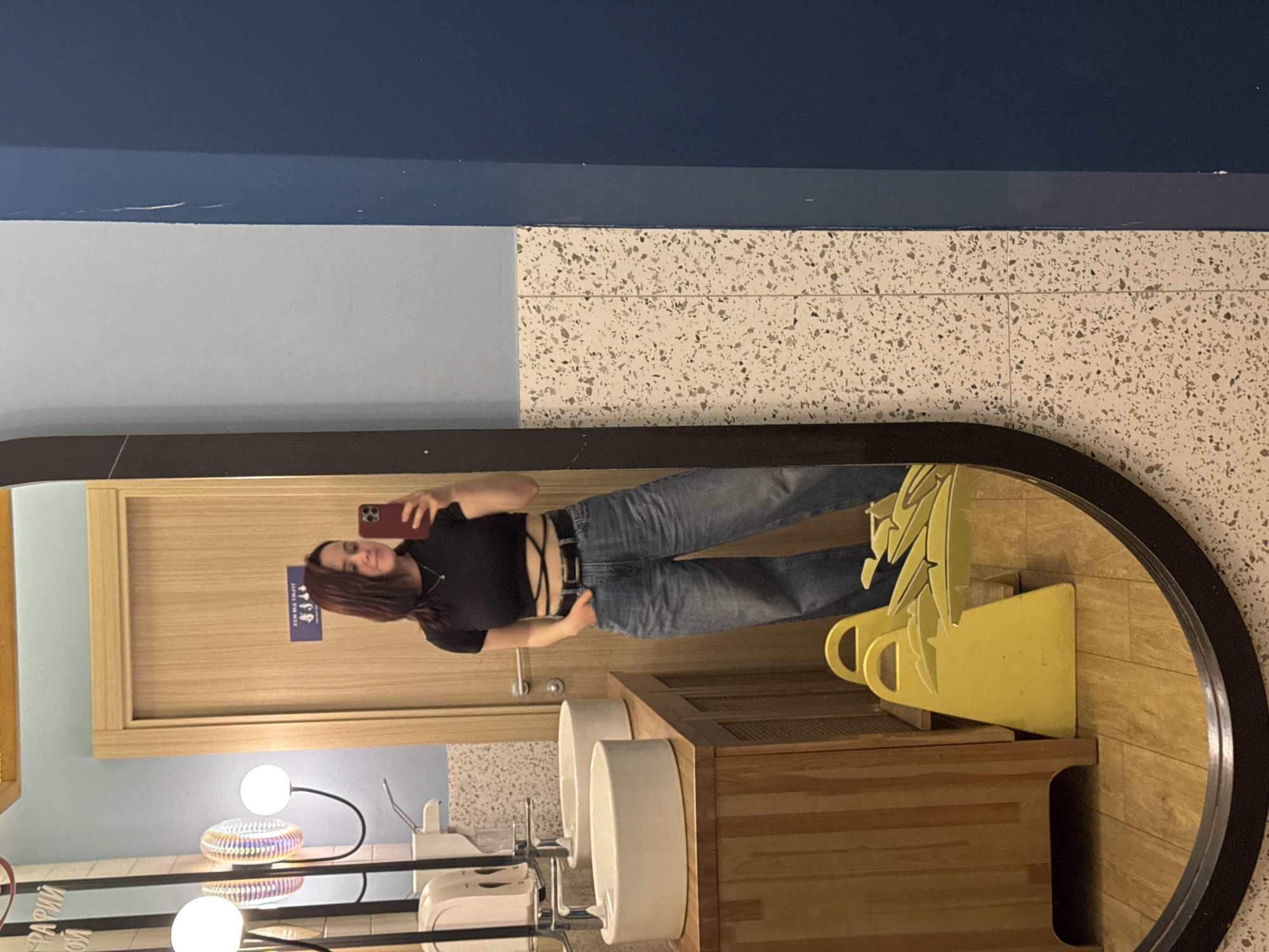 A woman stands in a public restroom taking a selfie in a large, circular mirror. The restroom features a blue wall divided into lighter and darker shades, with a section tiled in a speckled pattern. The woman is wearing a black top and blue jeans, holding a smartphone up to capture her reflection. The restroom also includes modern sinks and fixtures, a wooden cabinet, and decorative lighting. The location is Izhevsk, Russia.